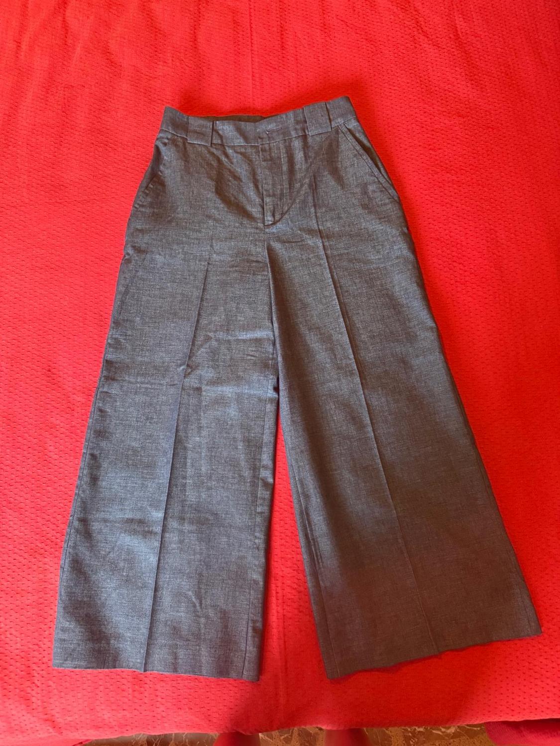 Grey wide-leg trousers with pleats and belt loops, laid out on vibrant red fabric. Relaxed composition with colour contrast.