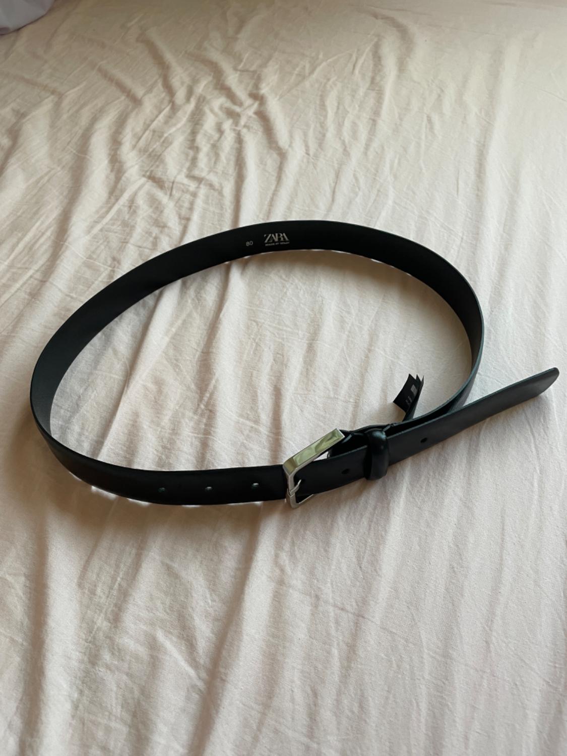 Black belt with a silver buckle on a white bed. Visible text: "ZARA 80".