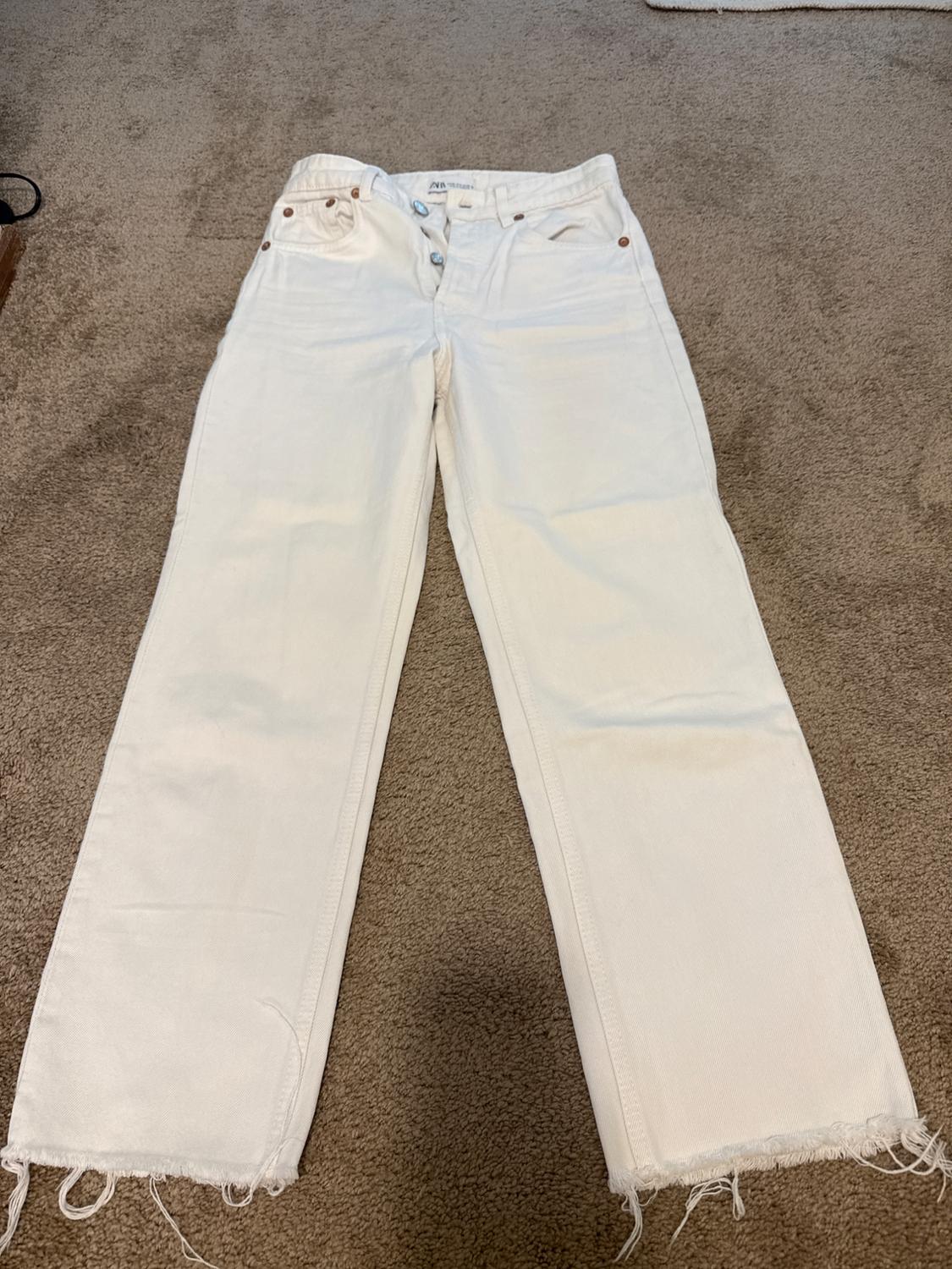 White ZARA jeans with a button closure and frayed hem, laid out on a brown rug.