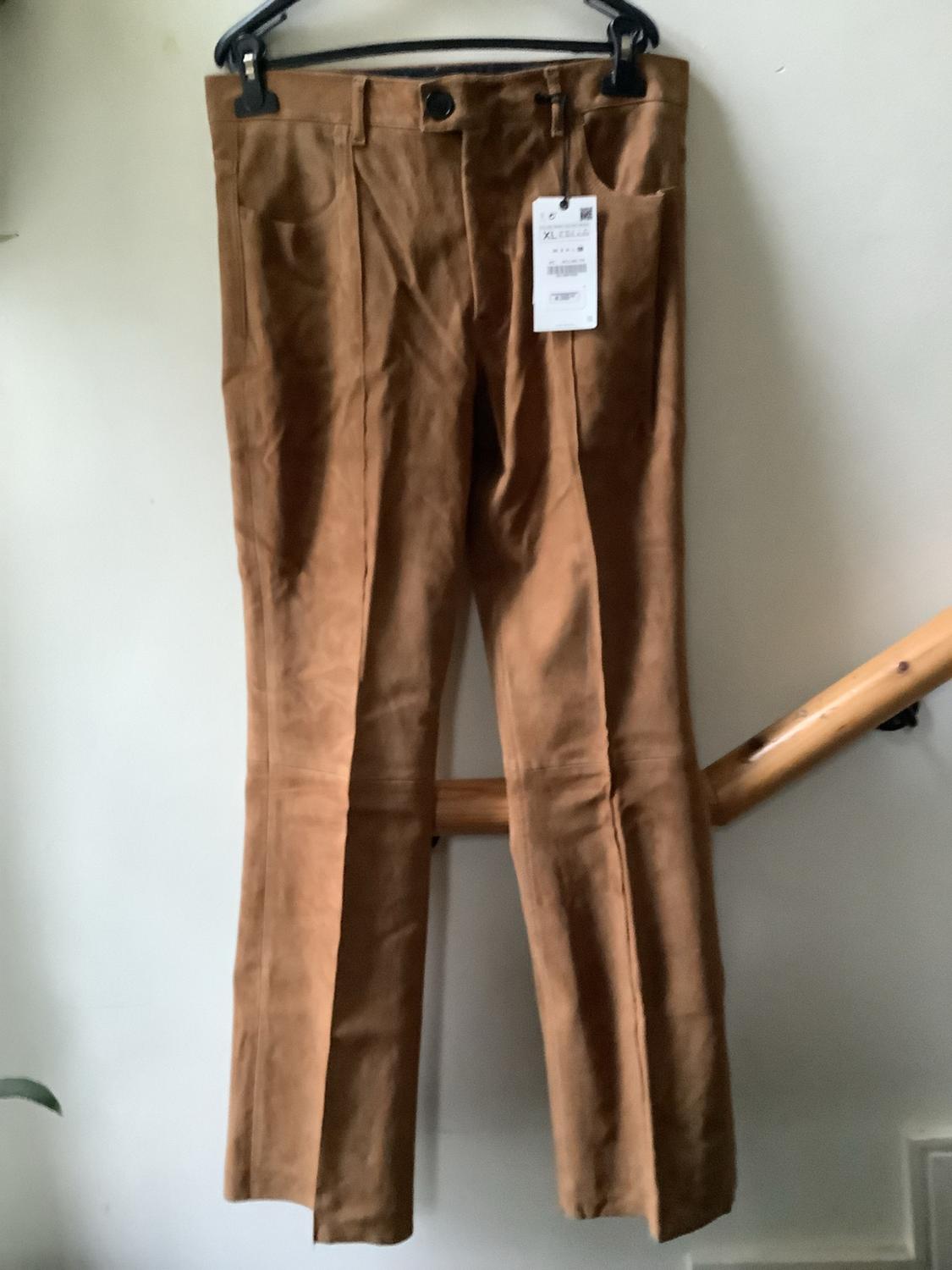 Brown trousers with a front seam hanging on a black hanger against a white wall, next to a wooden banister. Label with the text “XL”.
