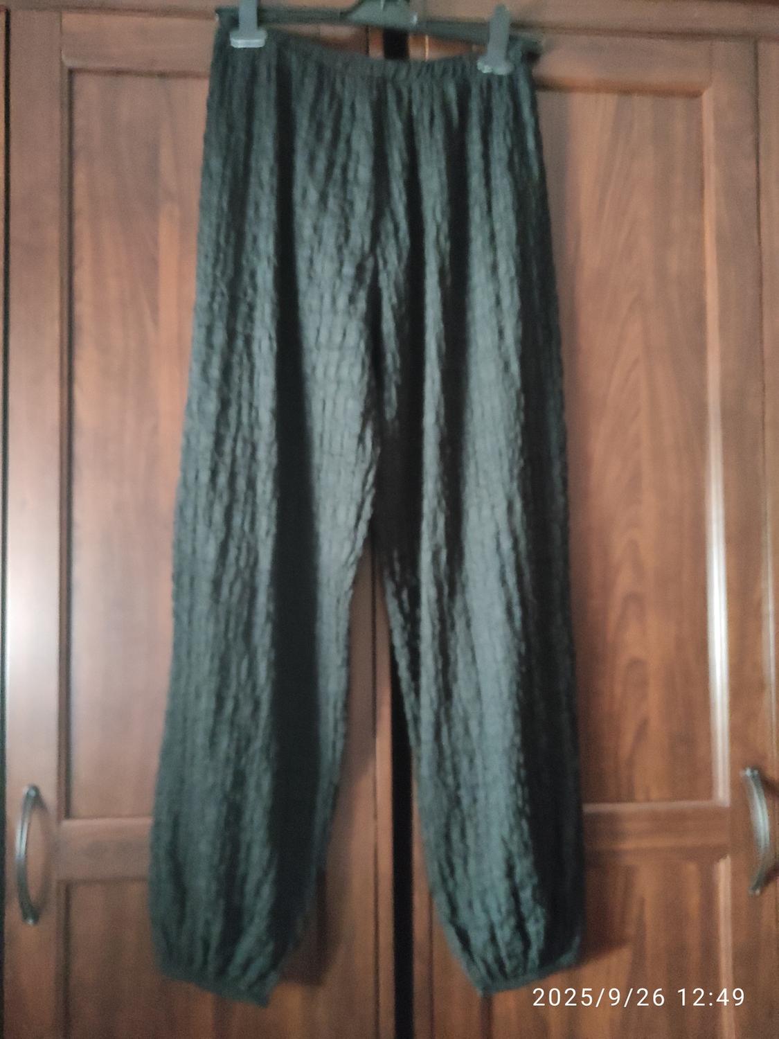 Dark trousers with a crinkled texture hanging on a hanger in front of a wooden wardrobe. Date in the corner: 2025/9/26 12:49.
