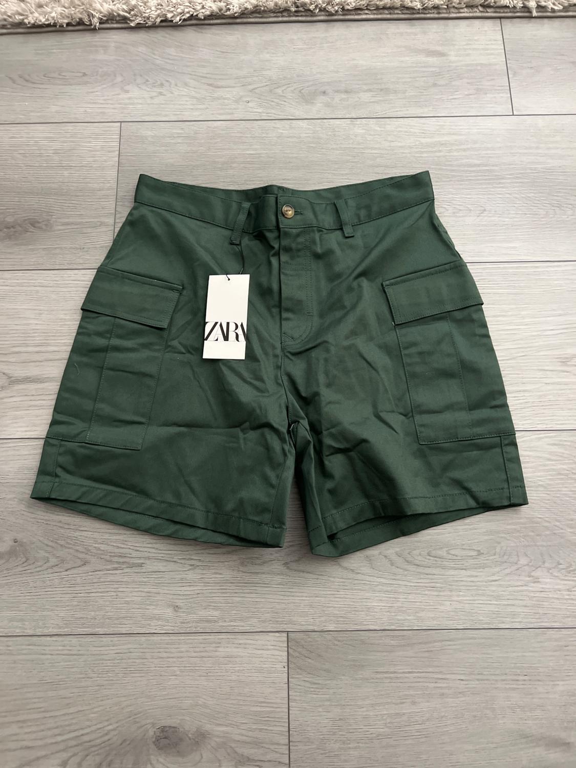 Dark green cargo shorts with side pockets, spread out on a gray wooden floor. Label with the text ZARA.