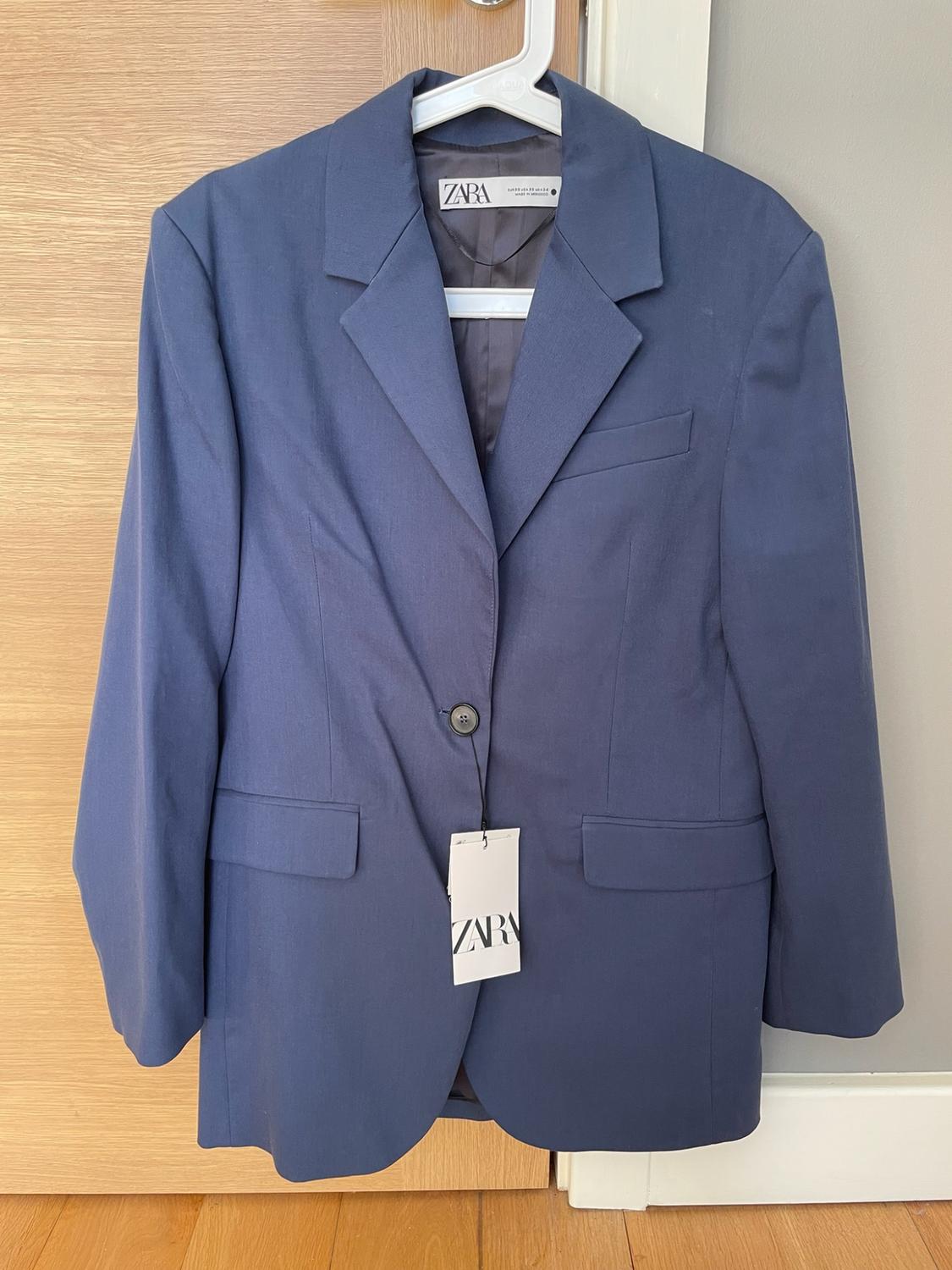 Navy blue blazer hanging on a white hanger over a wooden door. It shows a button fastened and labels with the text ZARA.