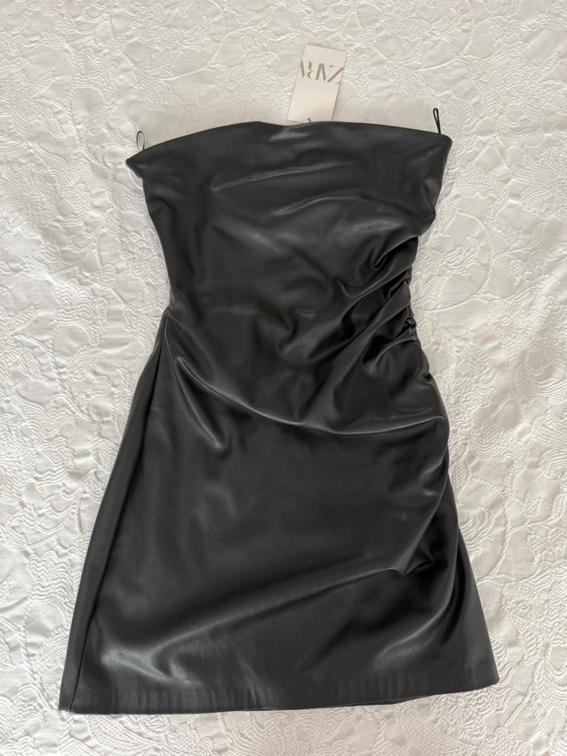Black strapless mini dress, with a shiny, draped finish, over textured white fabric. Visible label with ZARA text.