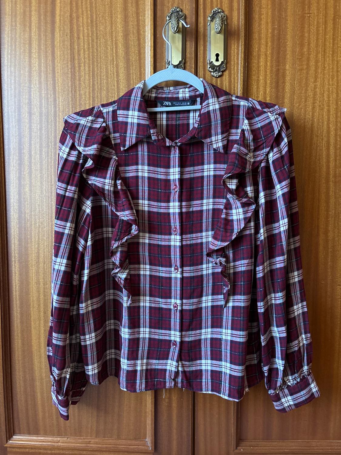 Red and white checked shirt with frills on the shoulders, hanging on a hanger in front of a wooden door.