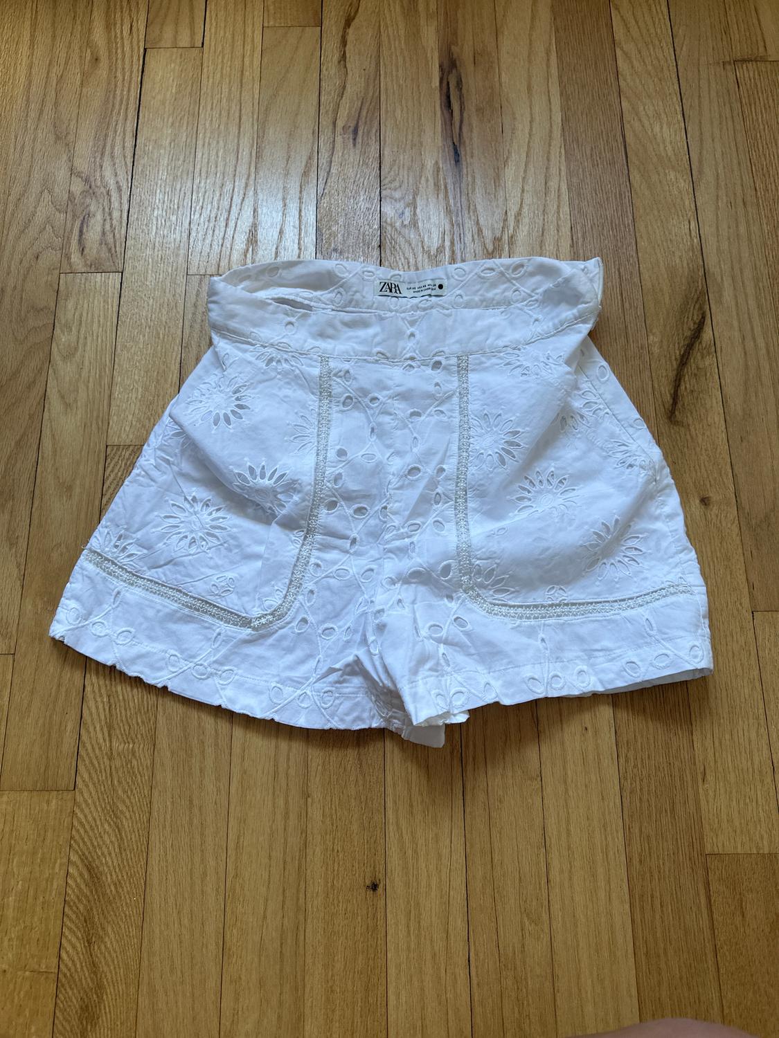 White lace shorts with a floral design on a wooden floor. Visible tag at the waist.