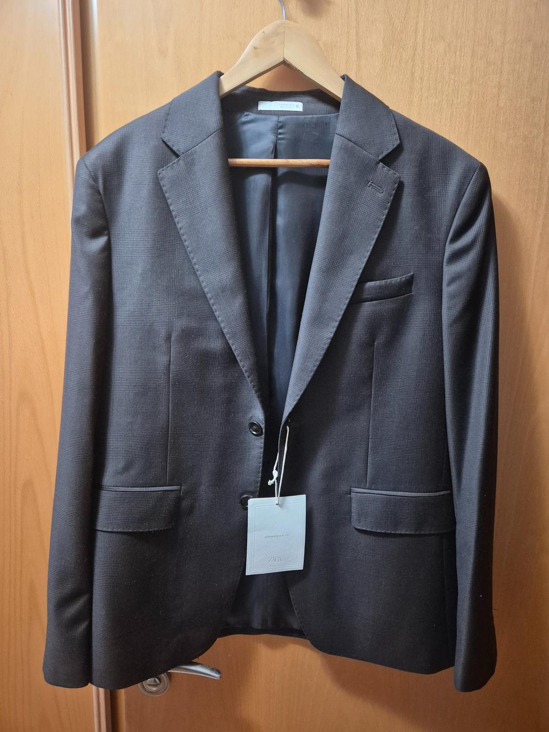 Black suit jacket hanging on a wooden hanger over a wooden door. Visible label with text "ZARA".