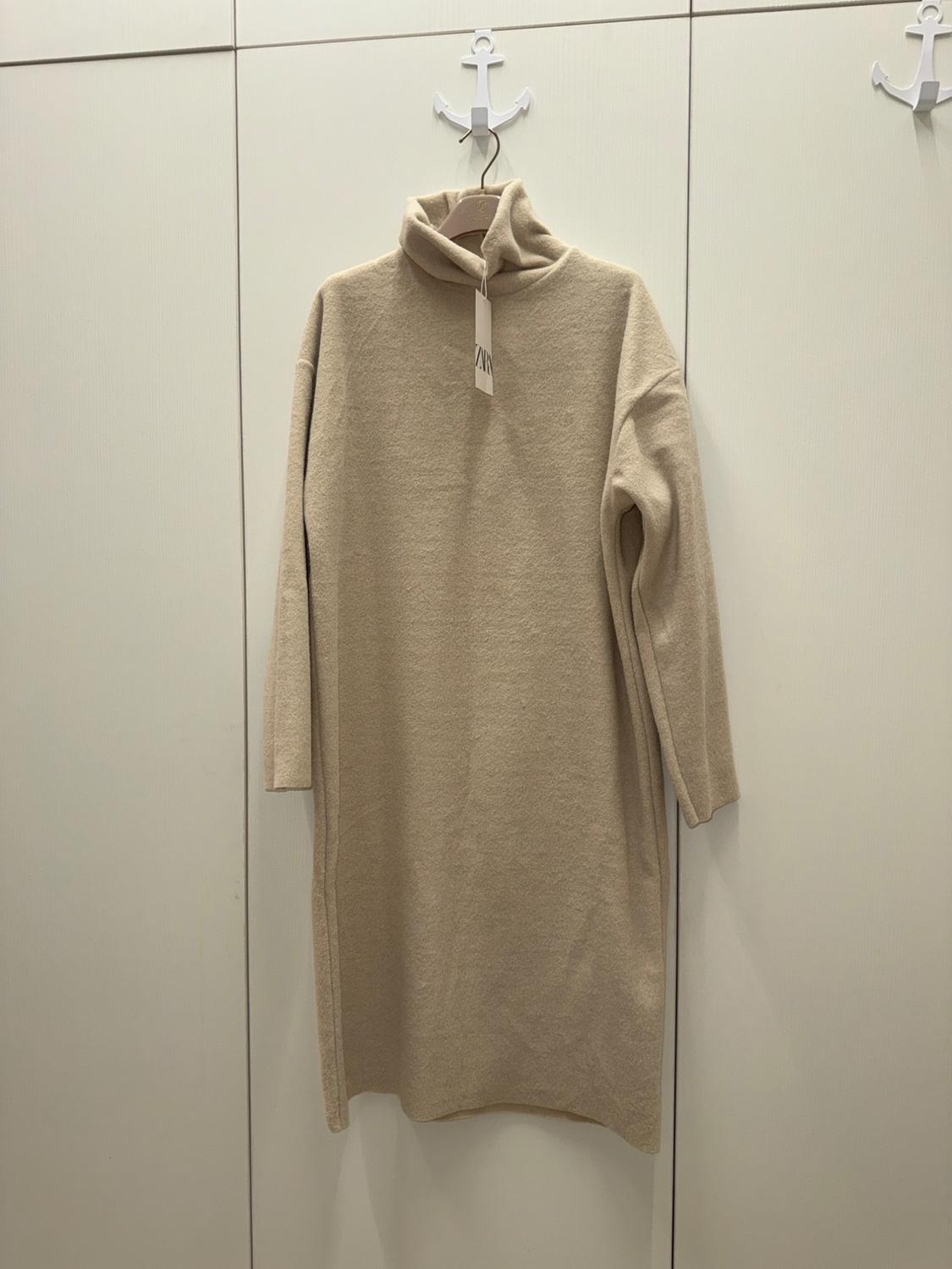 Beige long-sleeved, high-necked dress hanging on an anchor-shaped hanger against a white wall background.