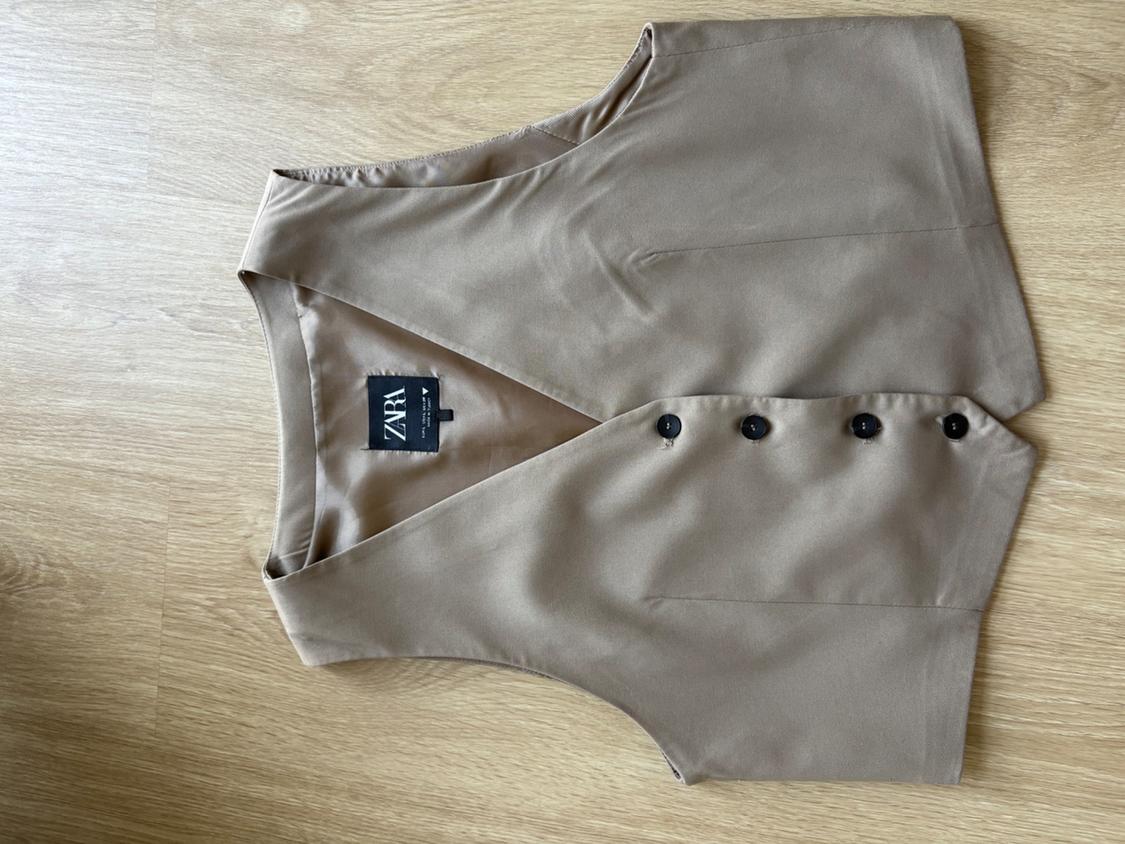 Beige tailored waistcoat with dark buttons on a light wooden floor. Inside label with the text ZARA.