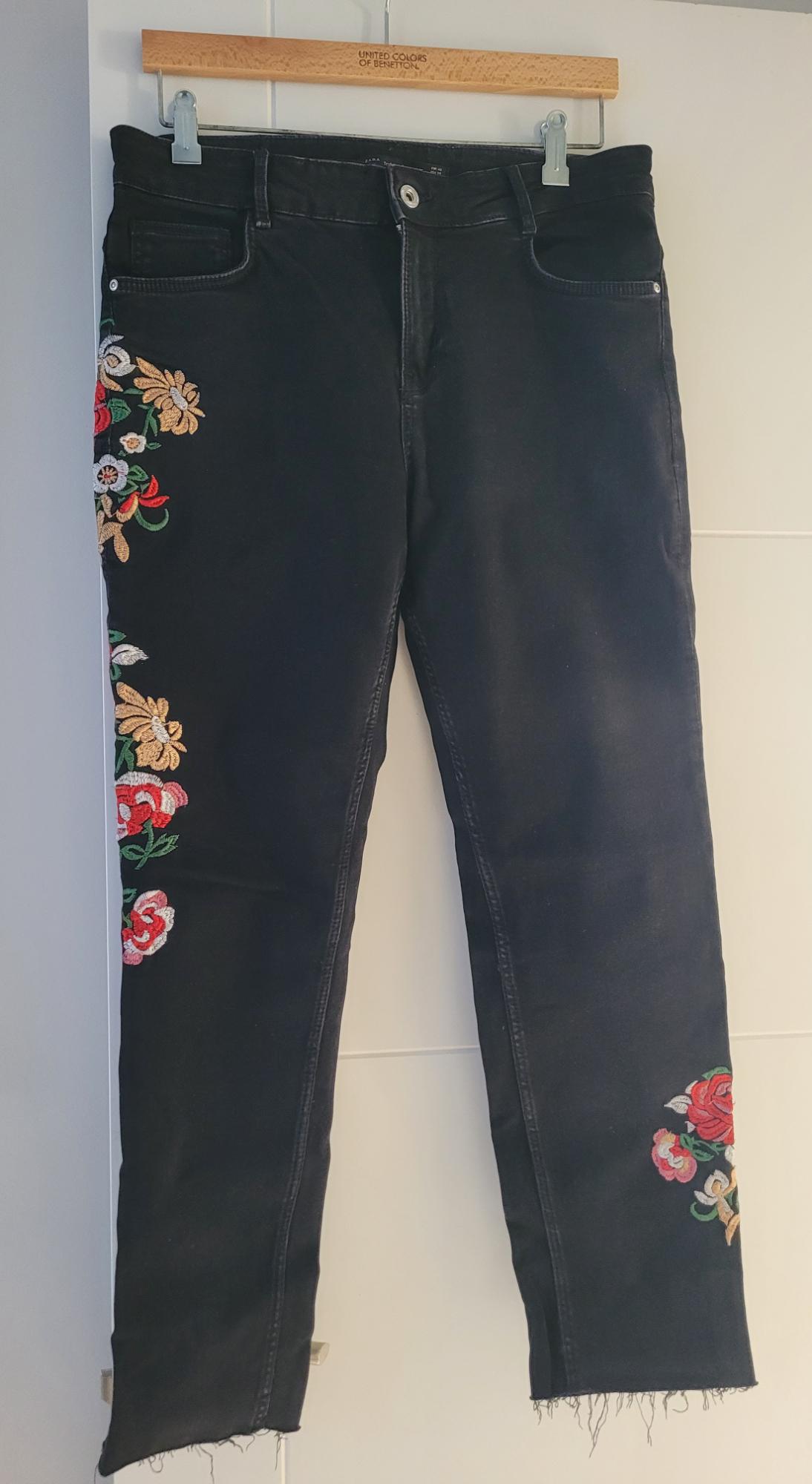 Black trousers with floral embroidery on the sides, hanging on a wooden hanger with "United Colors of Benetton" text.
