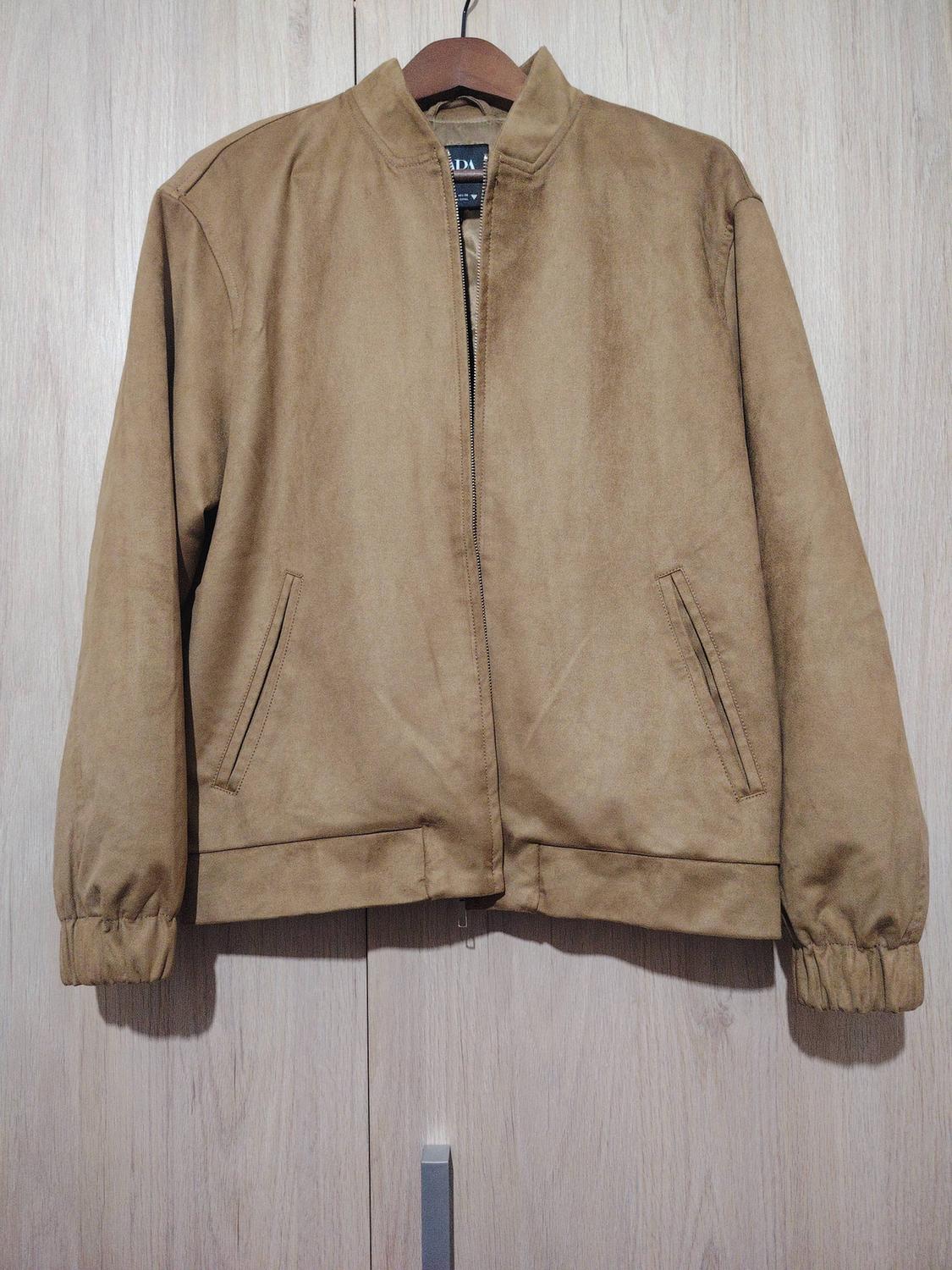 Light brown suede jacket hanging on a wooden hanger, with a front zip and side pockets. Light wood background.