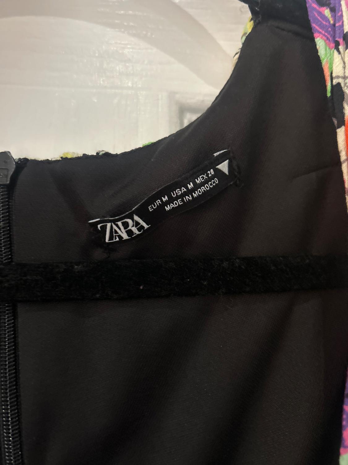 Black clothing label with text: "ZARA, EUR M USA M MEX 28, MADE IN MOROCCO". Colourful fabric background with visible zip.