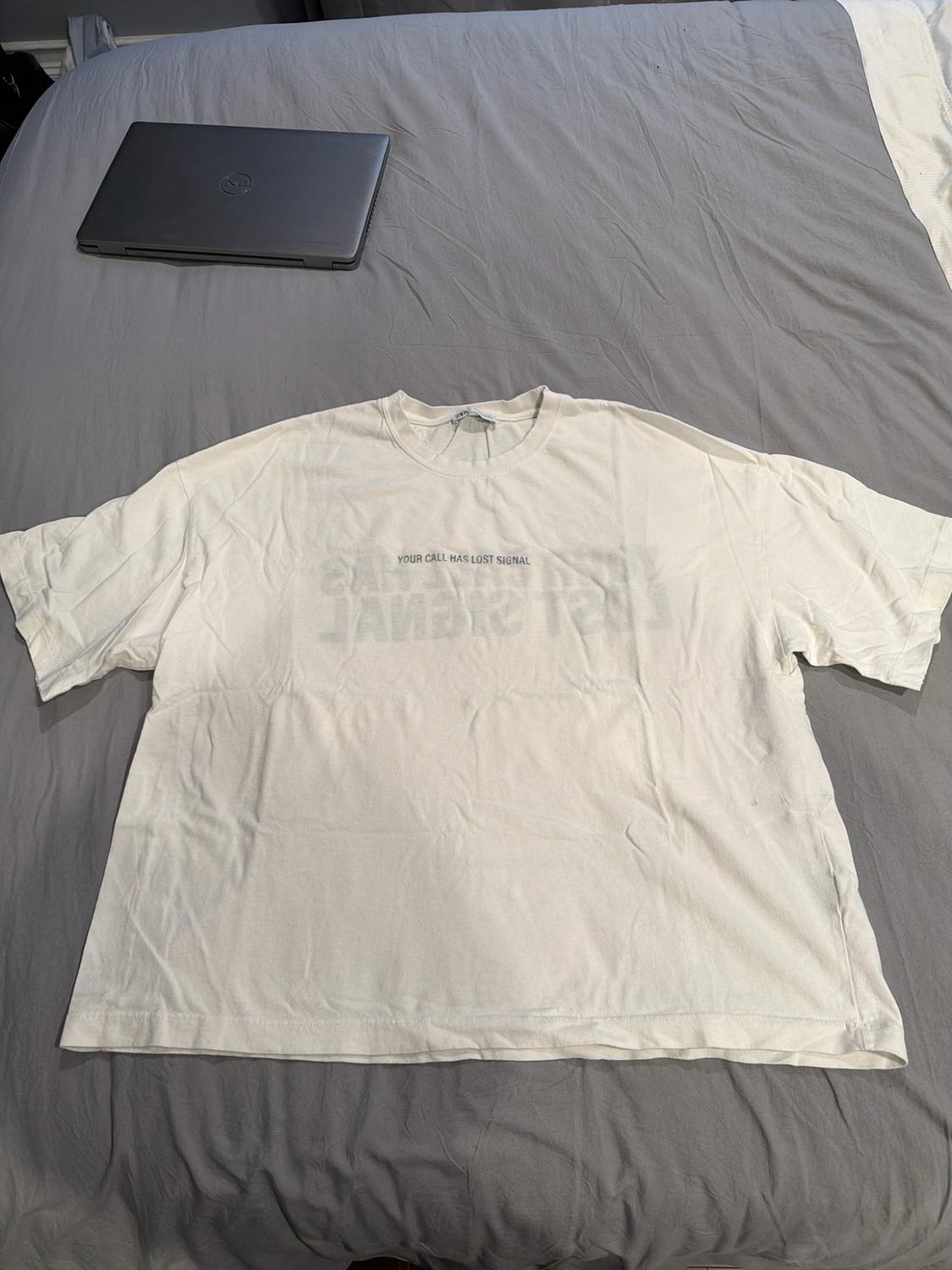 White short-sleeved T-shirt on a grey bed with a laptop. Casual style. Text: YOUR CALL HAS LOST SIGNAL.