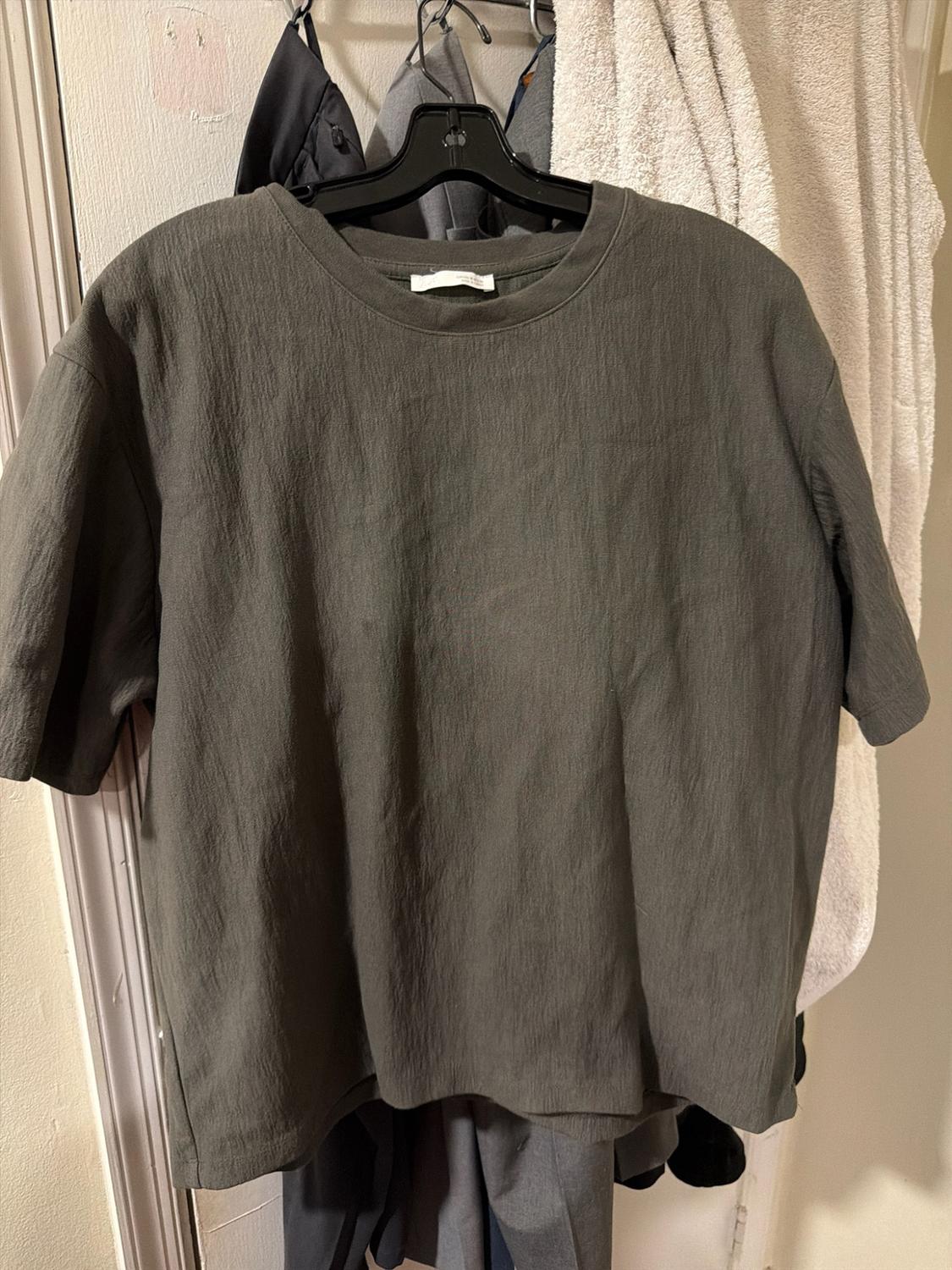 Dark grey short-sleeved t-shirt with a crinkled texture, hanging on a black hanger. In the background, other garments and a white towel.
