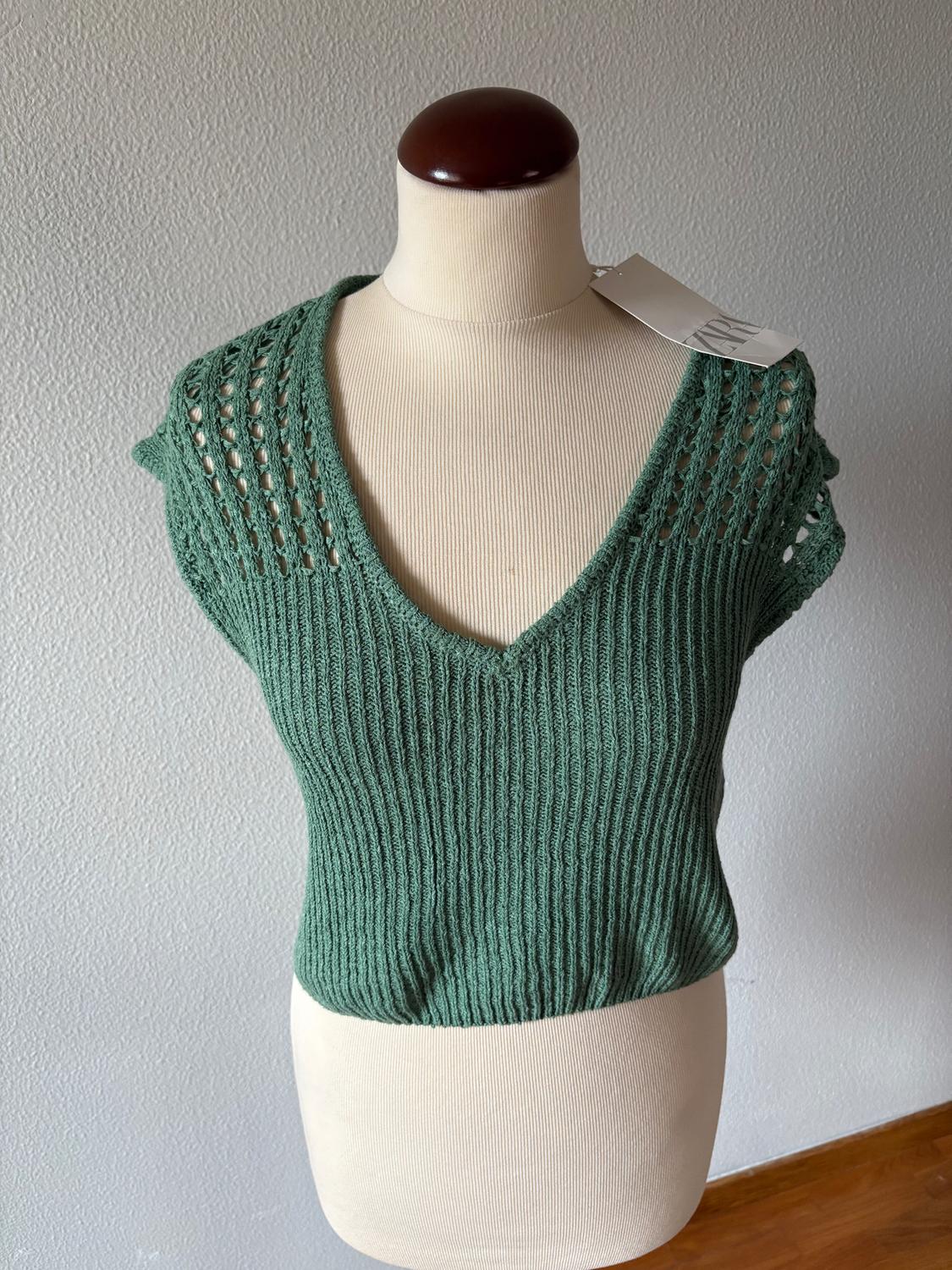 Mannequin in a green knitted V-neck top with openwork sleeves. White tag hanging from the right shoulder. Light grey background.