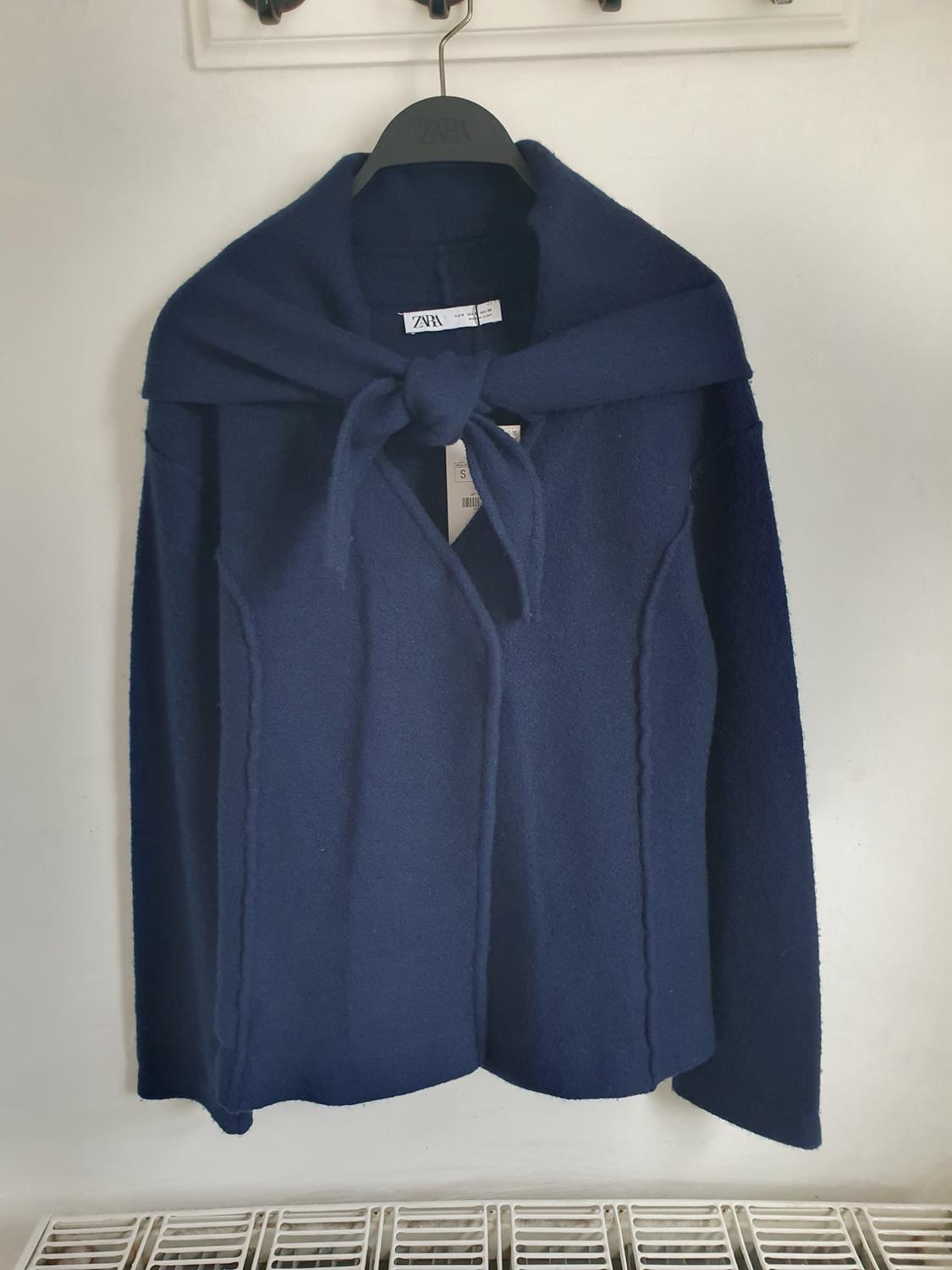Dark blue coat hanging on a black hanger with a visible label. White background and grid at the bottom of the image.