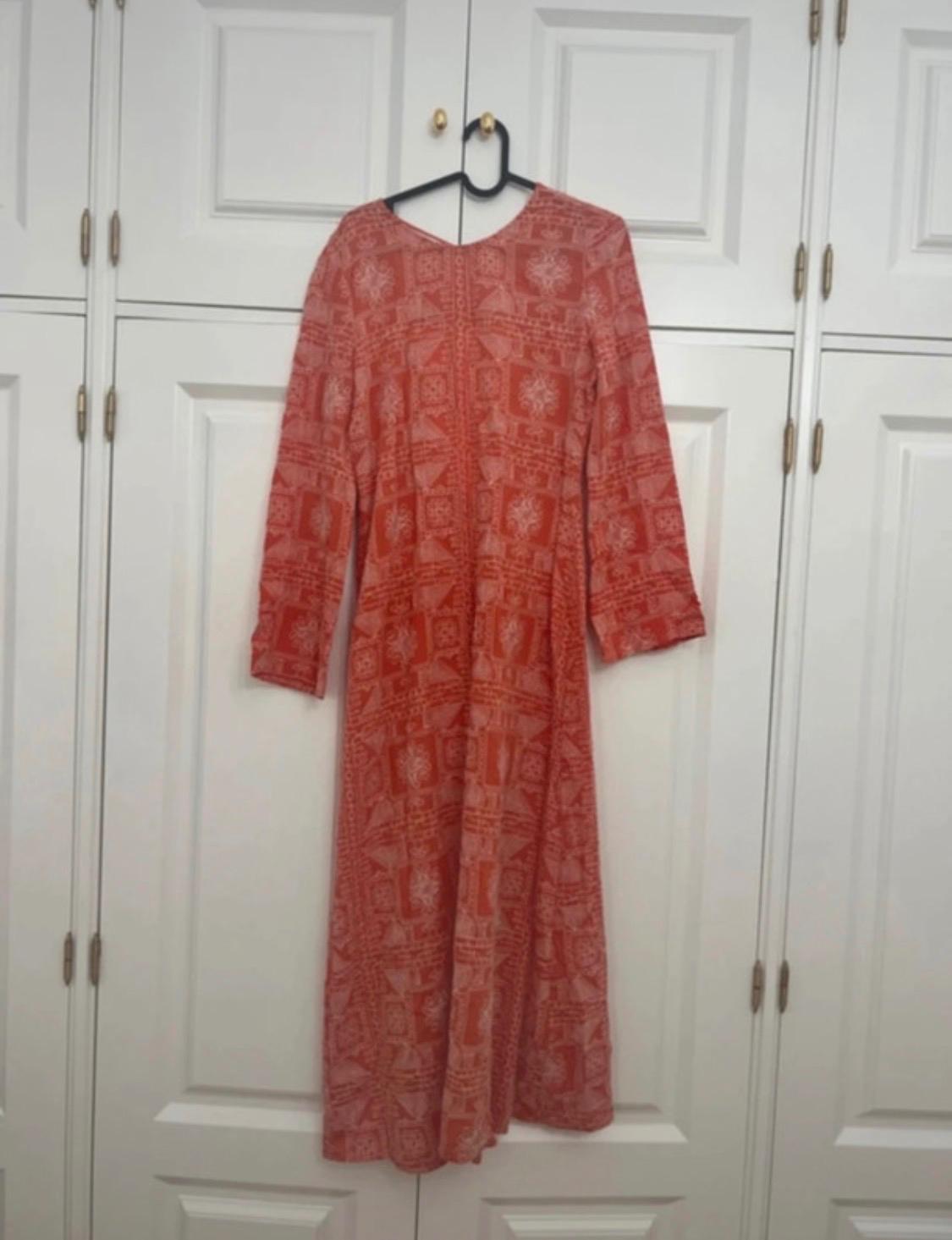 Long orange dress with a white print and long sleeves, hanging on a black hanger in front of a white wardrobe. Relaxed atmosphere.