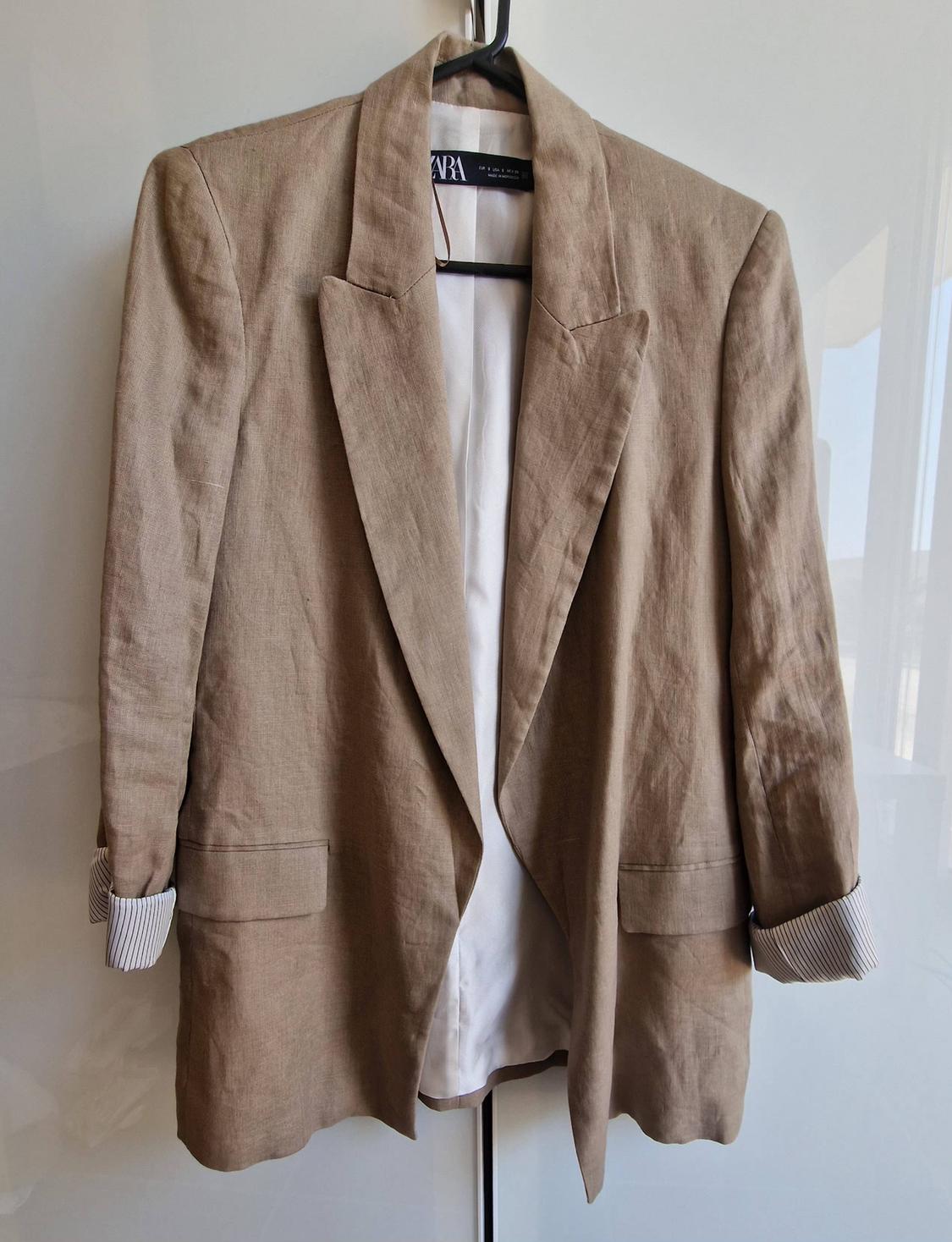 Beige jacket hanging on a black hanger against a white background. Rolled-up striped cuffs, flap pockets and white lining. Label with the text ZARA.
