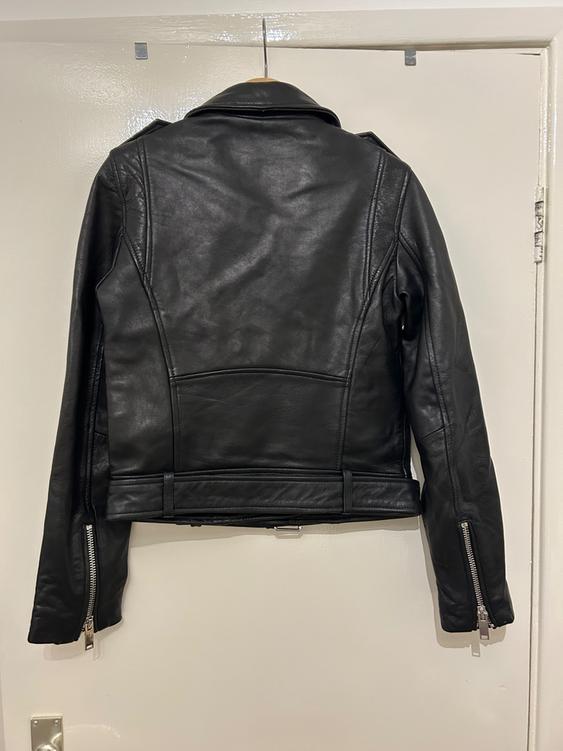 Black leather jacket hanging on a white door, viewed from behind. Zip details on the sleeves.