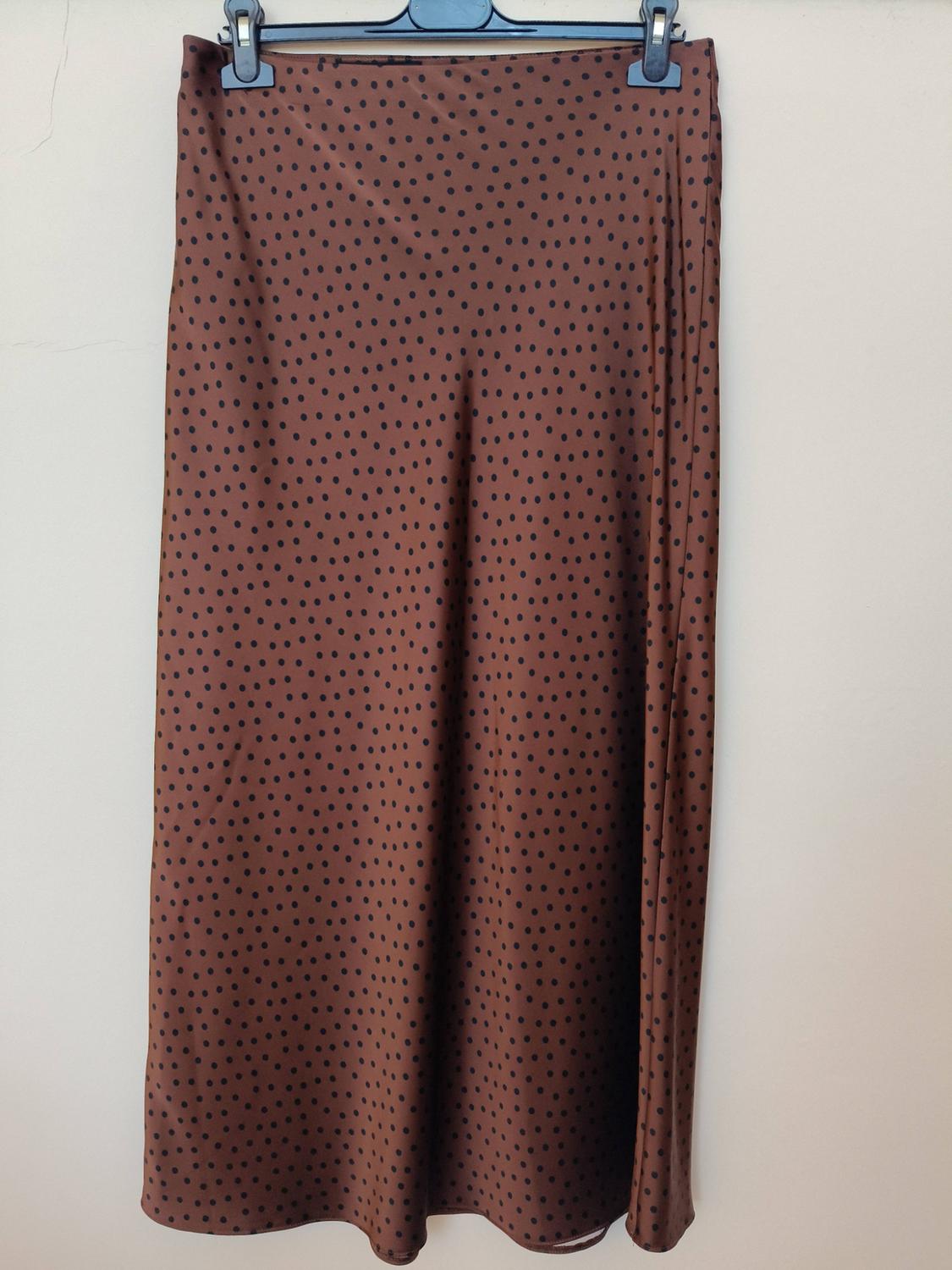 Brown skirt with a small black polka dot pattern, hanging on a black hanger in front of a light-coloured wall.