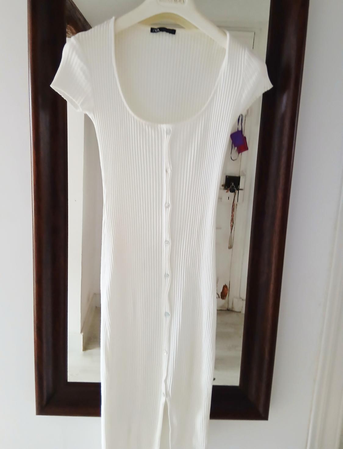 White ribbed dress, short sleeves and front buttons, hanging on a hanger in front of a dark wood-framed mirror.