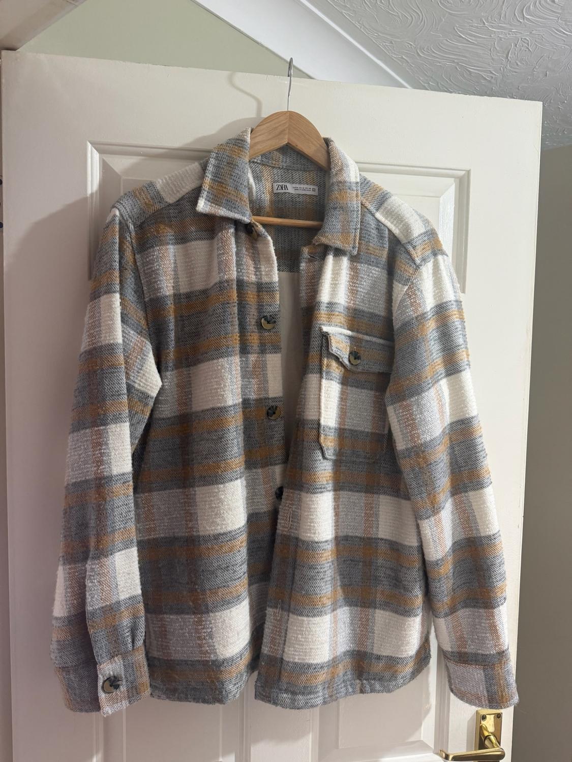 White, grey, and mustard check shirt with a ZARA label, hanging on a wooden hanger on a white door.