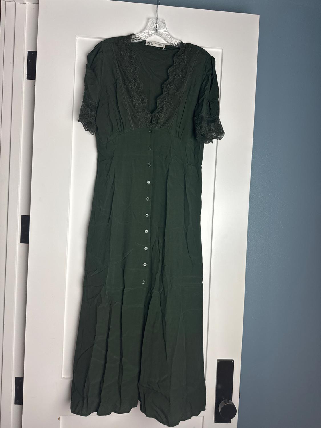 Dark green dress with front buttons and lace details on the neckline and short sleeves, hanging on a white door. Text: Zara.