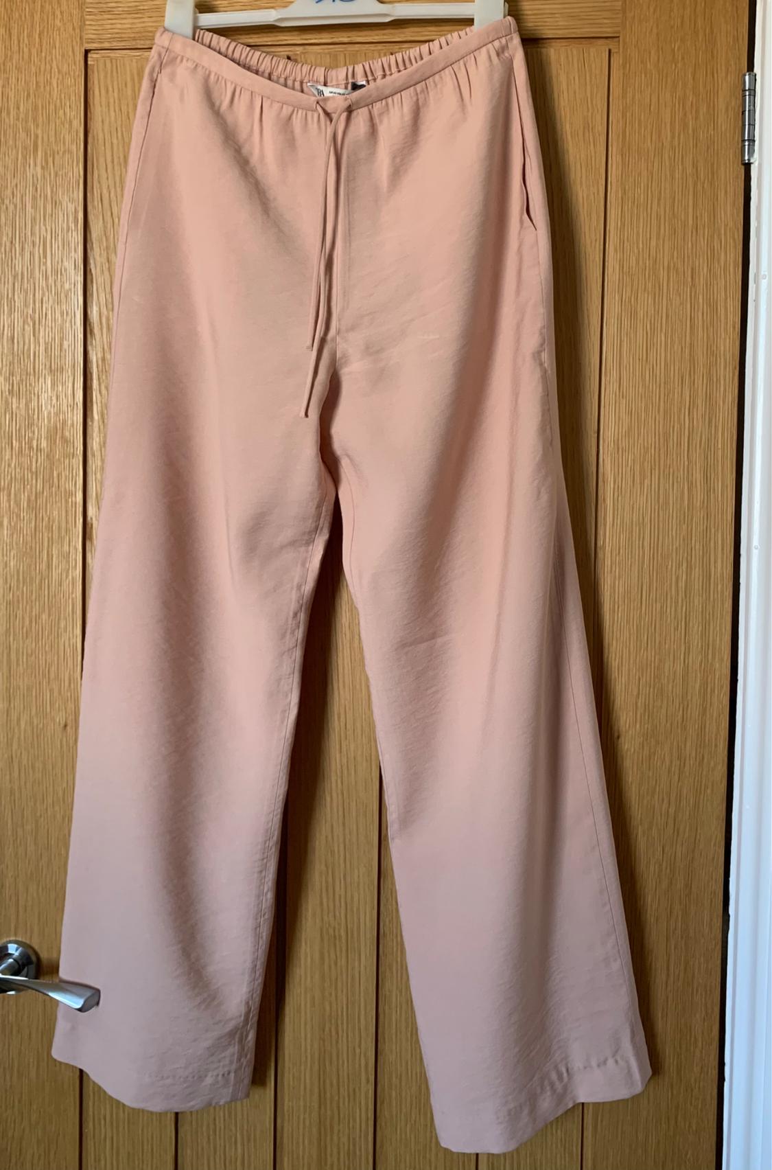 Light pink wide-leg trousers hanging on a wooden door, with an elasticated waistband and adjustable drawstring.