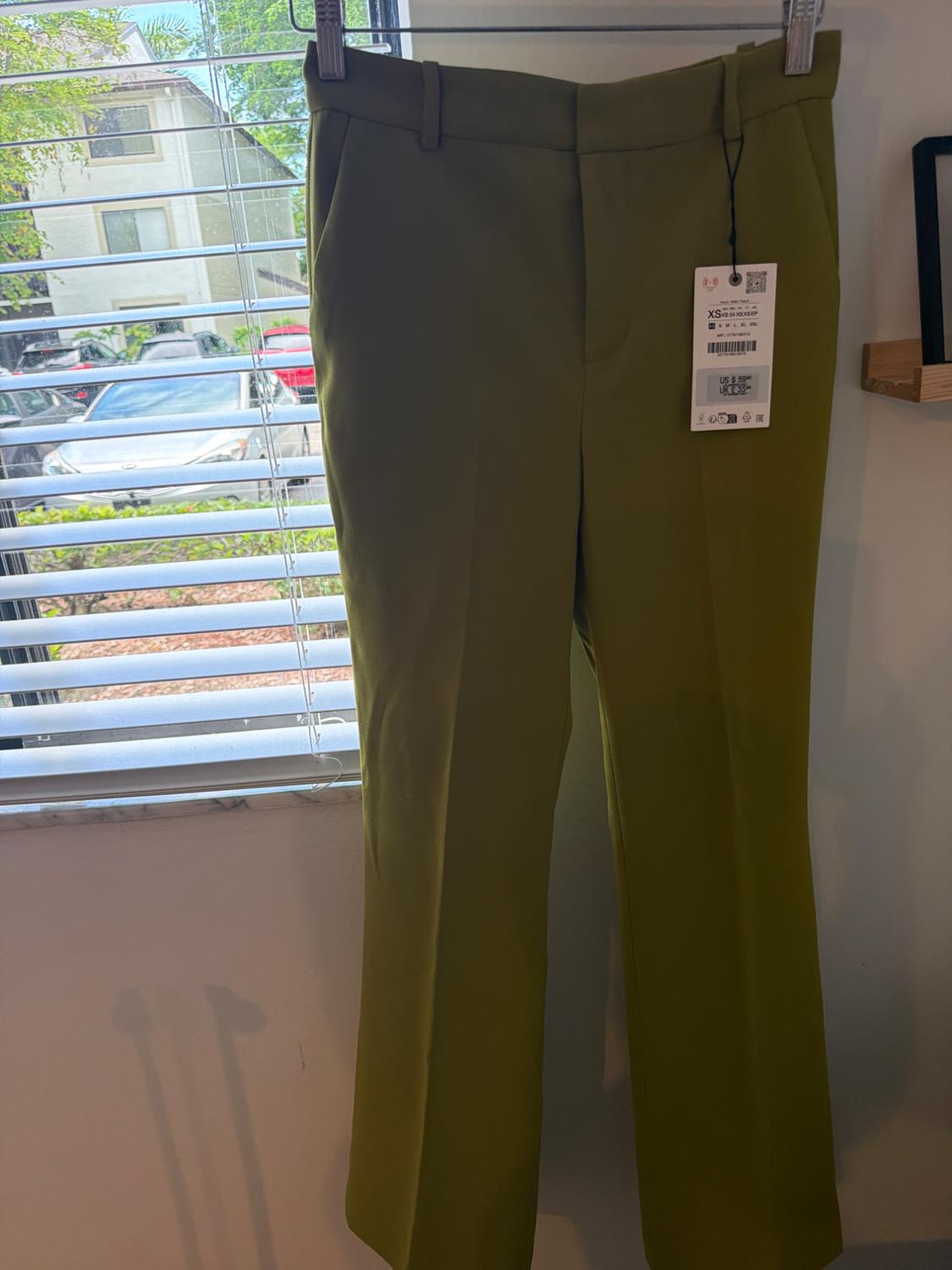 Olive green dress pants hanging in front of a window with blinds. Tag with text: XS US $ 59.90 UK £ 35.99.