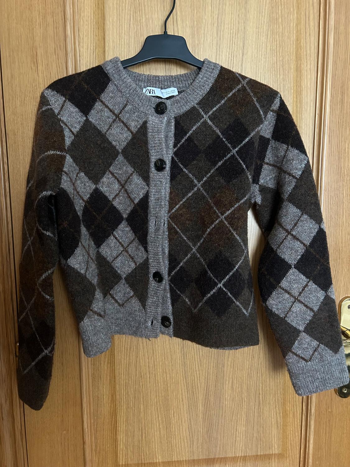 Wool cardigan with a diamond pattern in brown and grey tones, hanging on a black hanger over a wooden door.