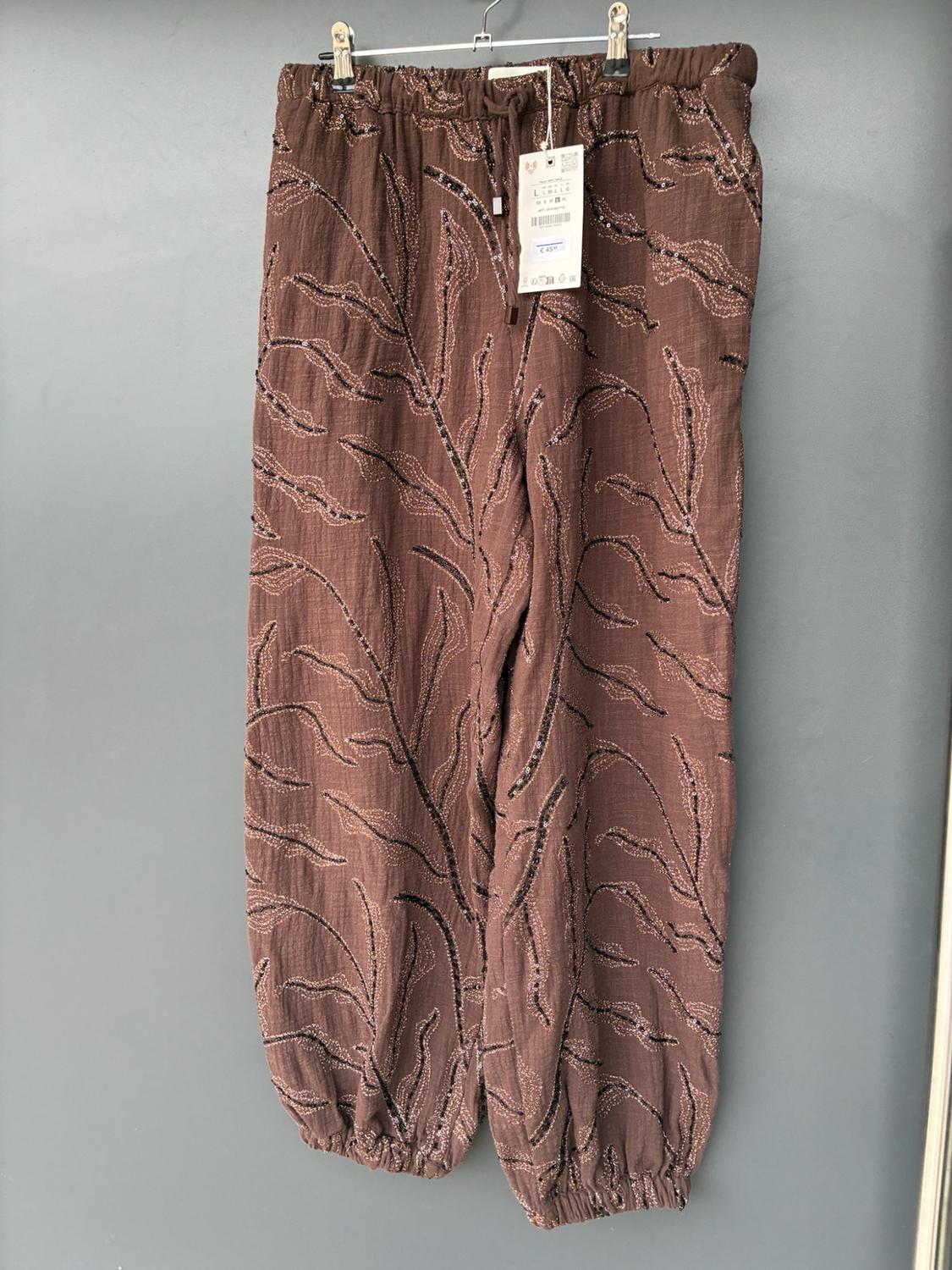 Loose brown trousers hanging on a hanger against a grey background. They have an elasticated waist and hems, and a branch pattern with shiny details.