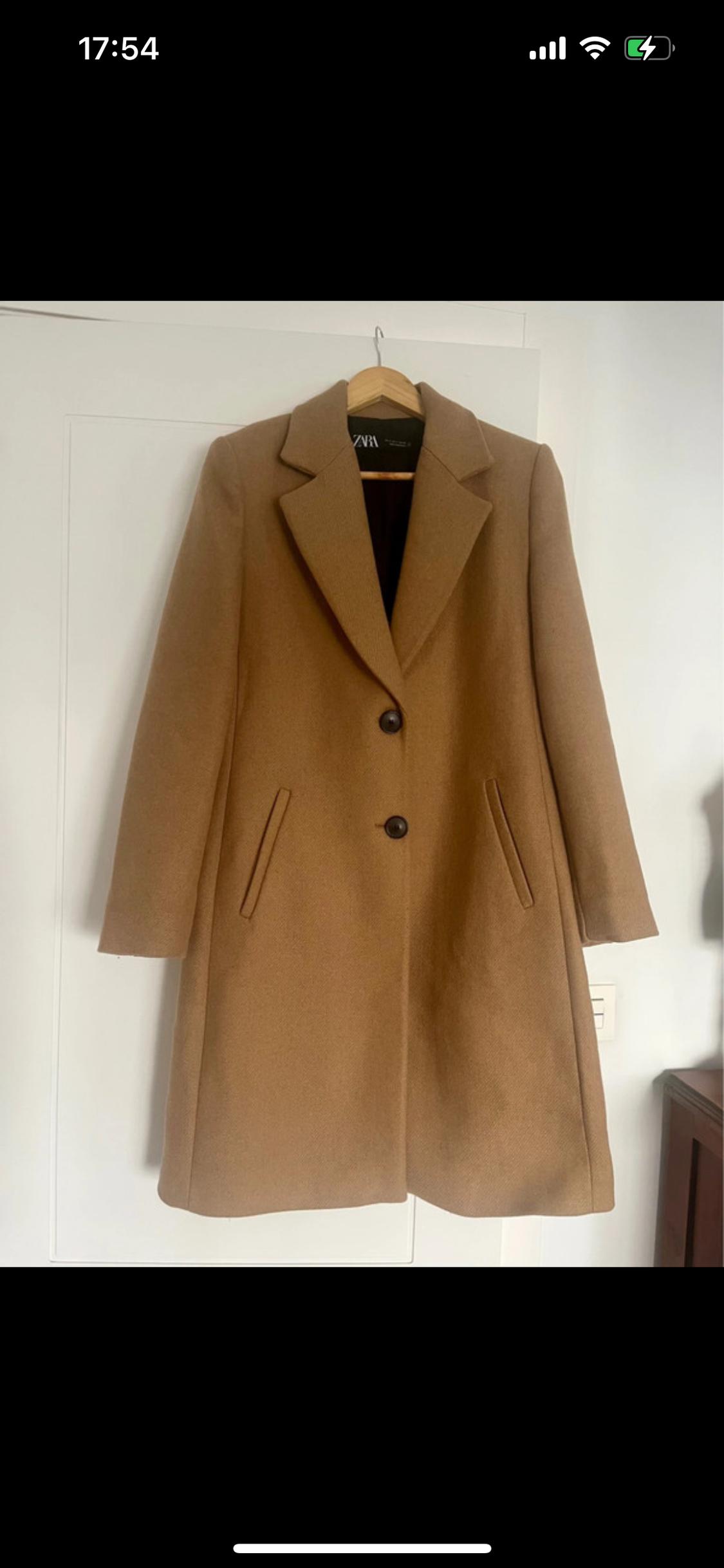 Camel coat hanging on a wooden hanger, with two dark buttons and slanted pockets. White background. Visible text: "ZARA".