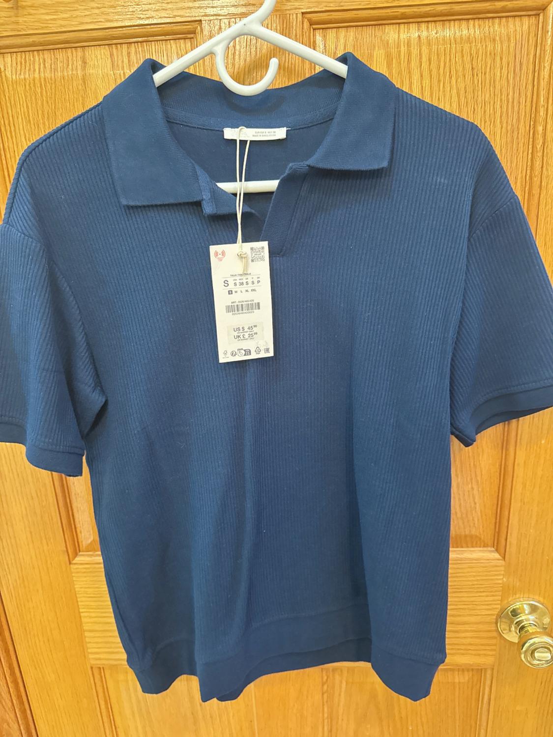 Blue short-sleeved polo shirt hanging on a white hanger over a wooden door. Visible tag with price of US$ 45.