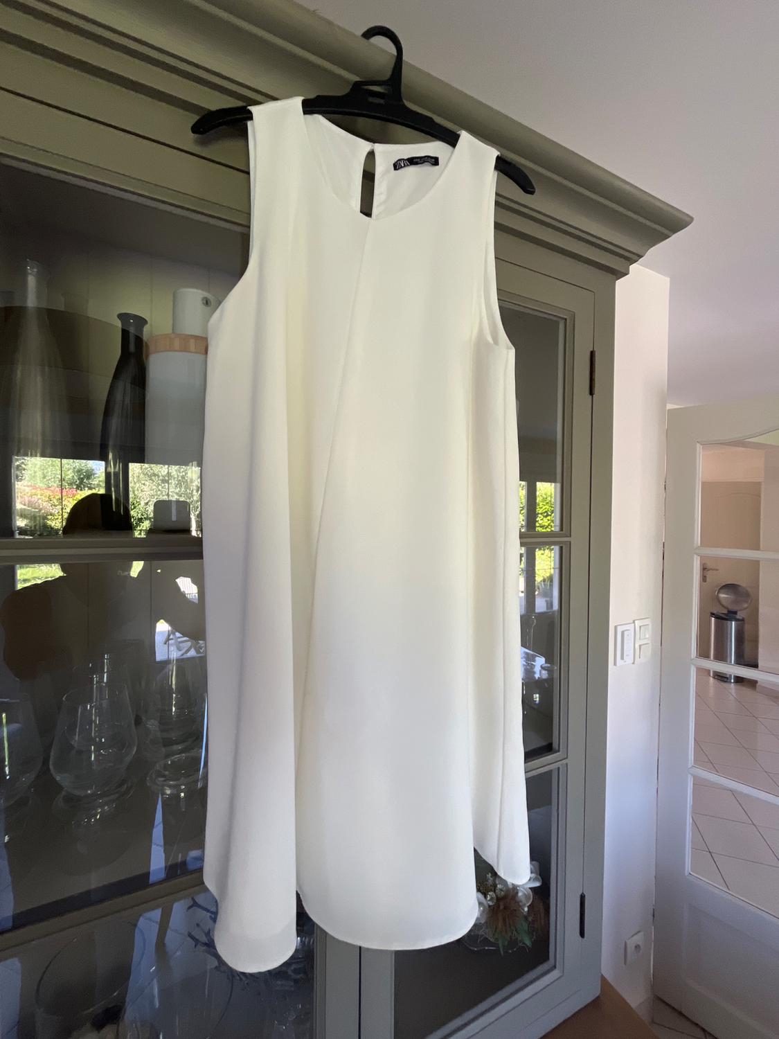 Flowing white sleeveless dress hanging on a black hanger above a grey display cabinet. Relaxed atmosphere in the bright interior of a house.