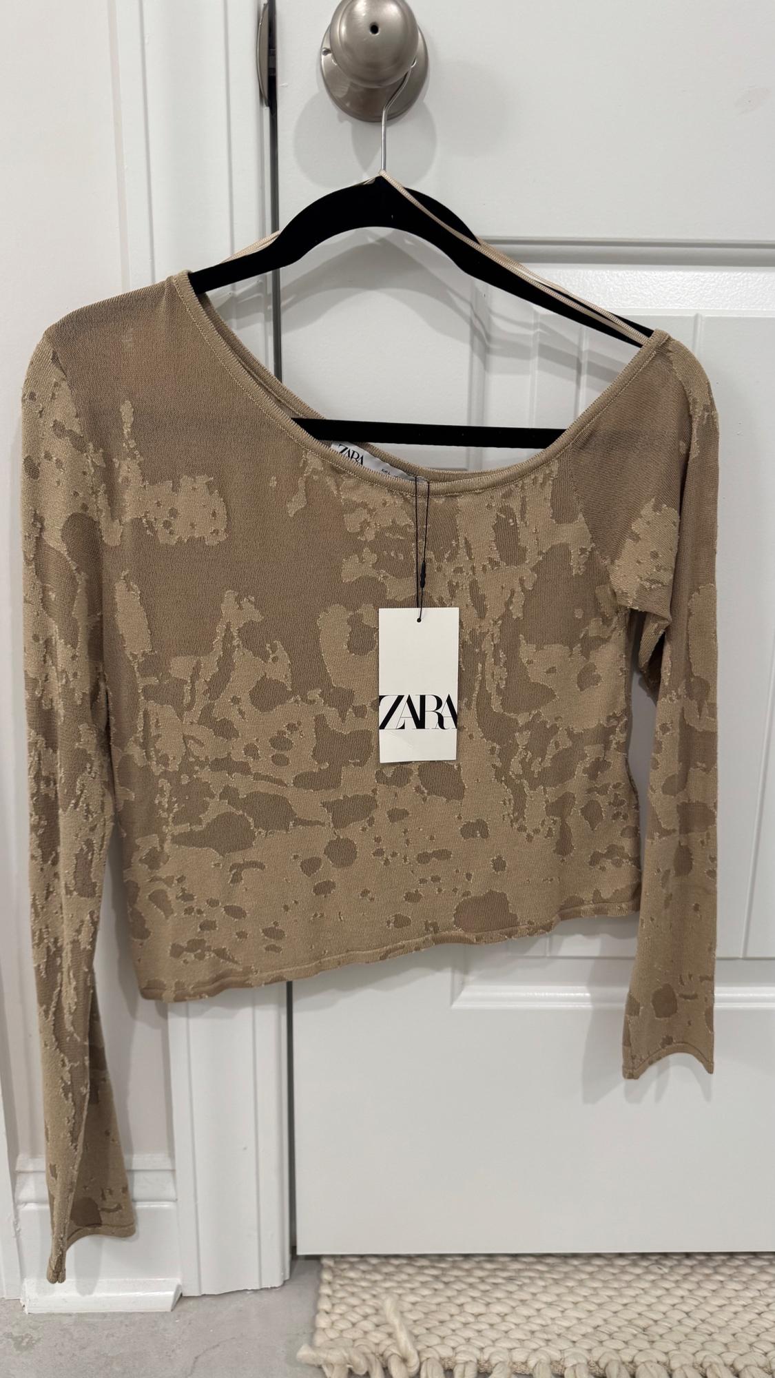 Beige jumper with an abstract design hanging on a black hanger in front of a white door. Label visible with the text "ZARA".