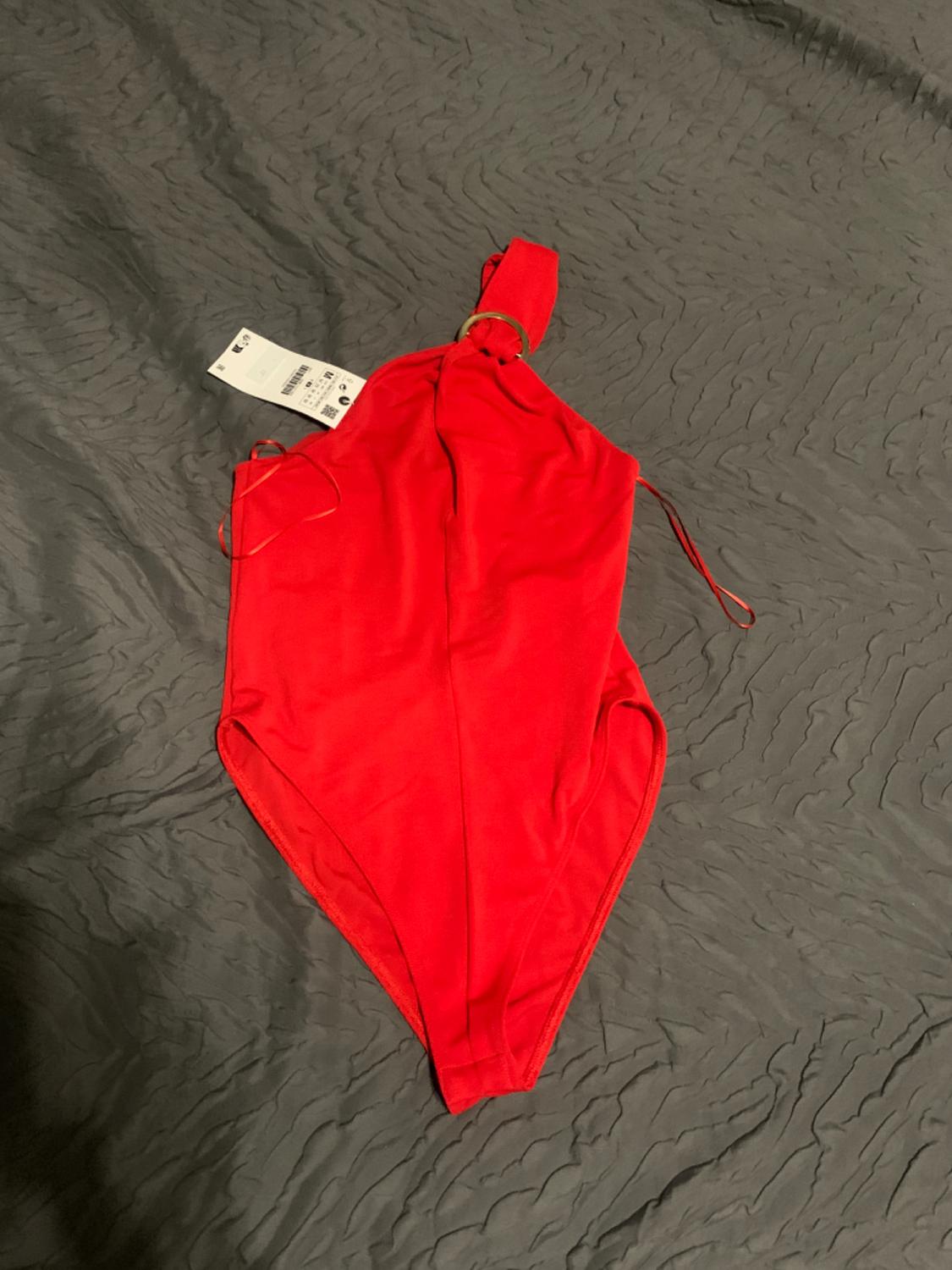 One-piece red swimming costume with tag, on a textured grey bedspread.