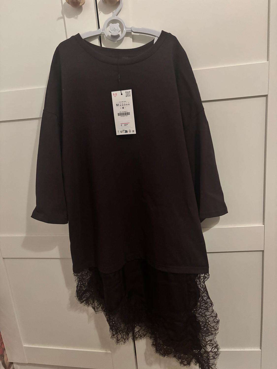 Μαύρο φόρεμα με δαντέλα στο στρίφωμα, κρεμασμένο σε μια λευκή κρεμάστρα μπροστά από μια λευκή ντουλάπα. Ορατή ετικέτα στο κέντρο.