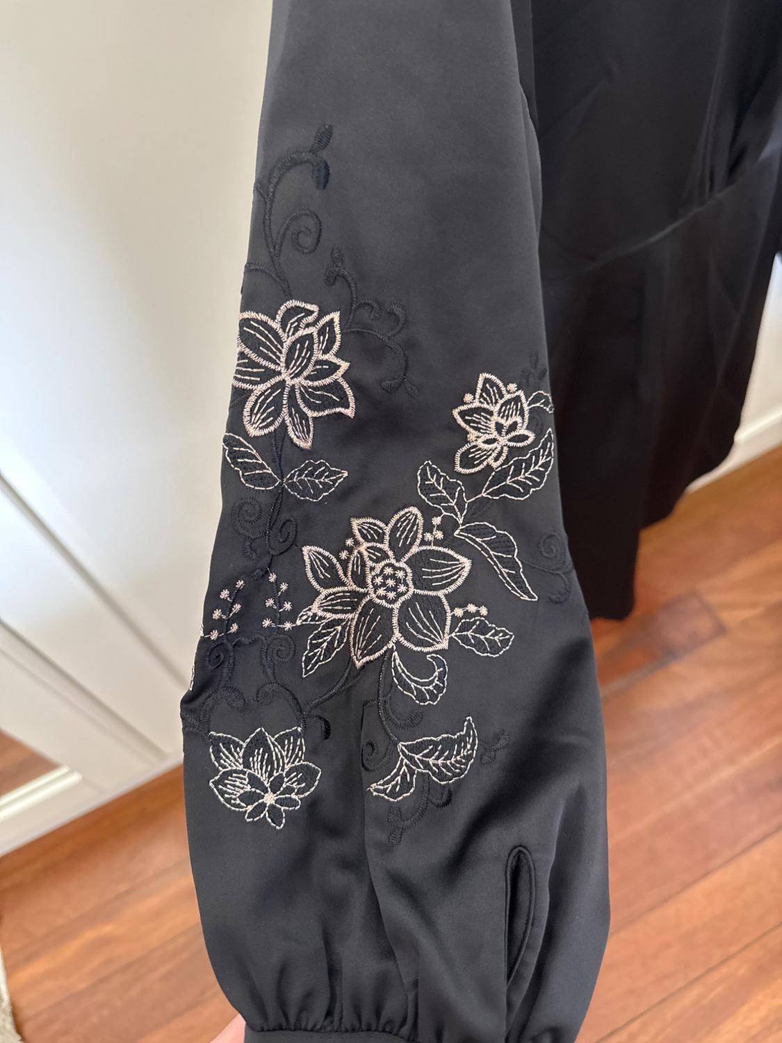 Close-up of a black sleeve with a gathered cuff and light floral embroidery. Relaxed indoor setting with a wooden floor.