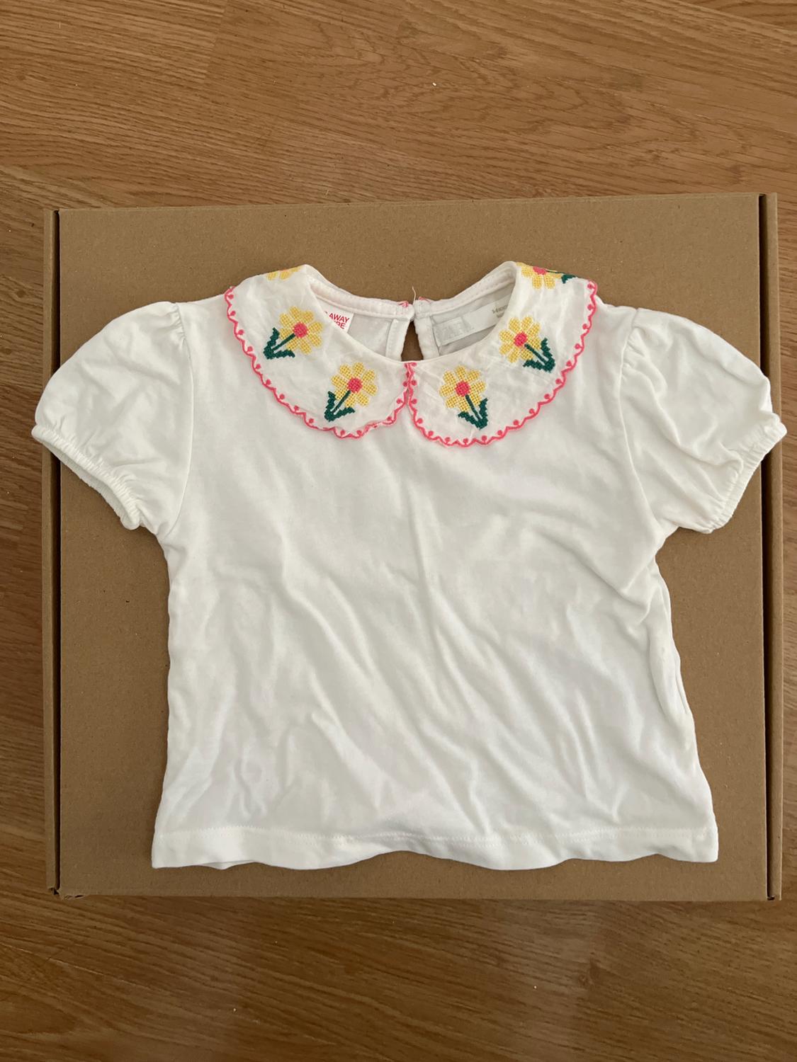 Short-sleeved white blouse with an embroidered collar featuring yellow flowers and pink details, on a brown cardboard background.