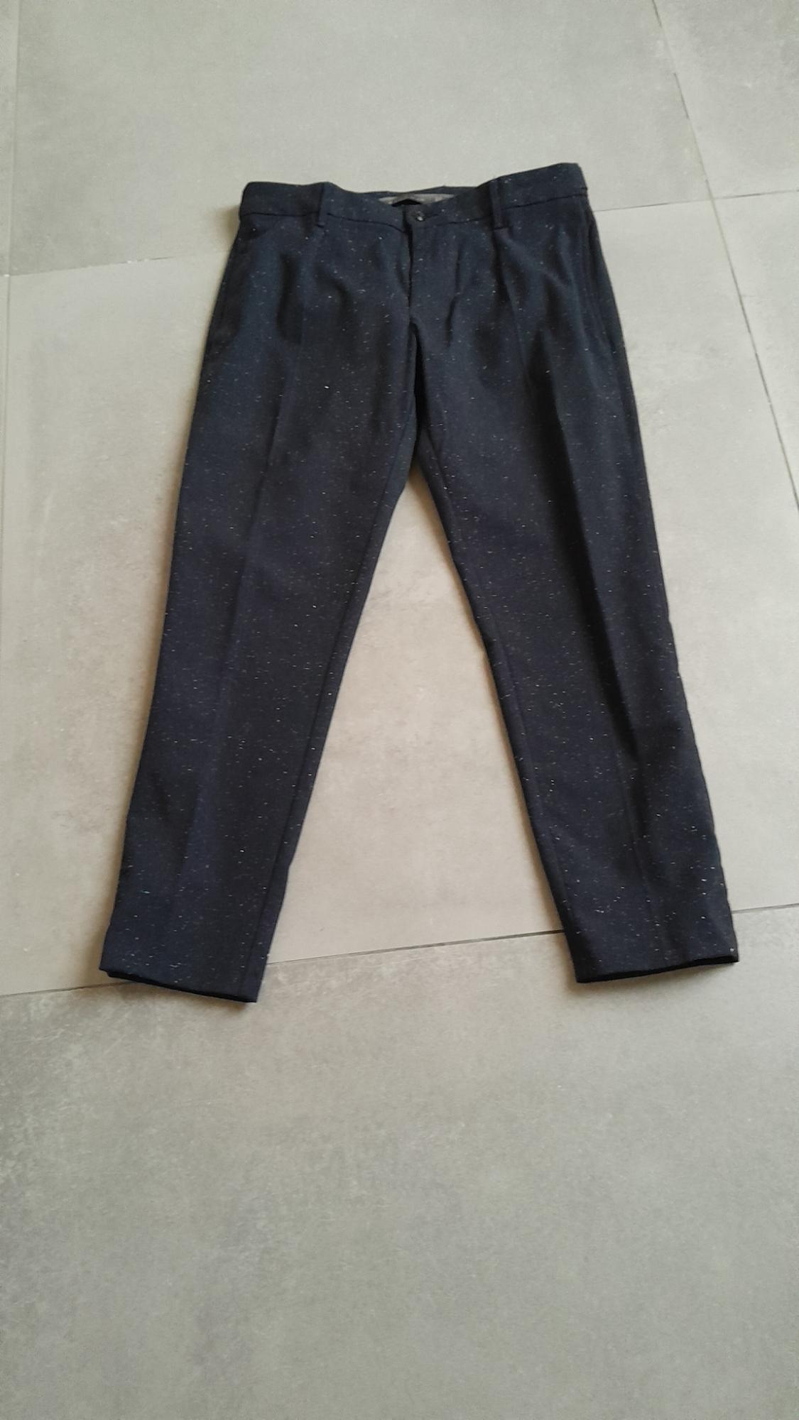 Dark trousers with a mottled texture, placed on a light-coloured floor. Classic design with pleats and button fastening.