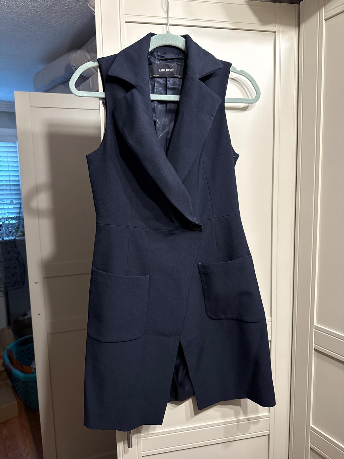 Navy blue sleeveless vest with large lapels and front pockets, hanging on a green hanger in a closet. Text: "ZARA BASIC".