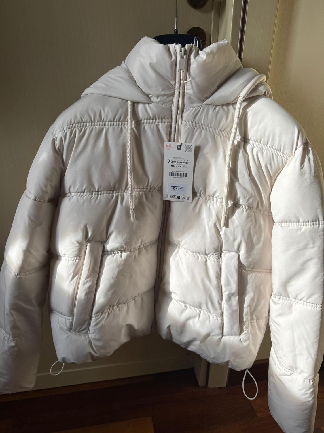 White padded jacket hanging on a hanger, with a hood and visible tag showing size XS and a price of €49.99.