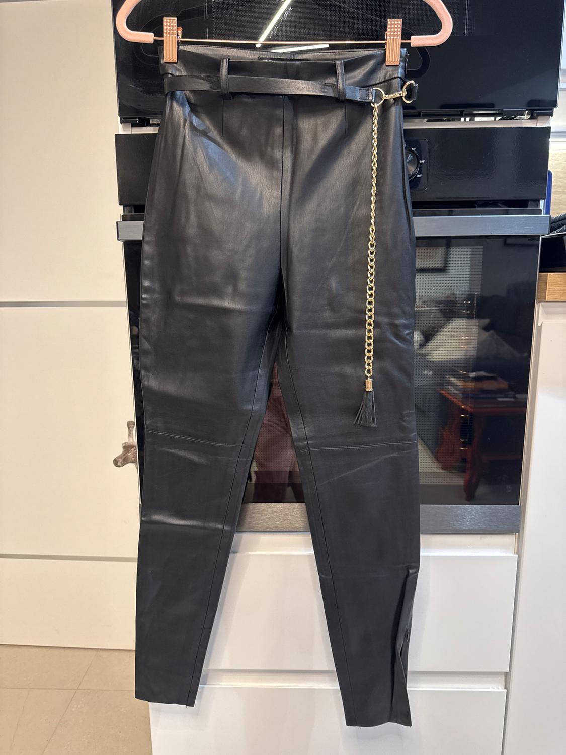 Black leather trousers hanging on a hanger, with a belt and decorative gold chain, against a modern kitchen background.