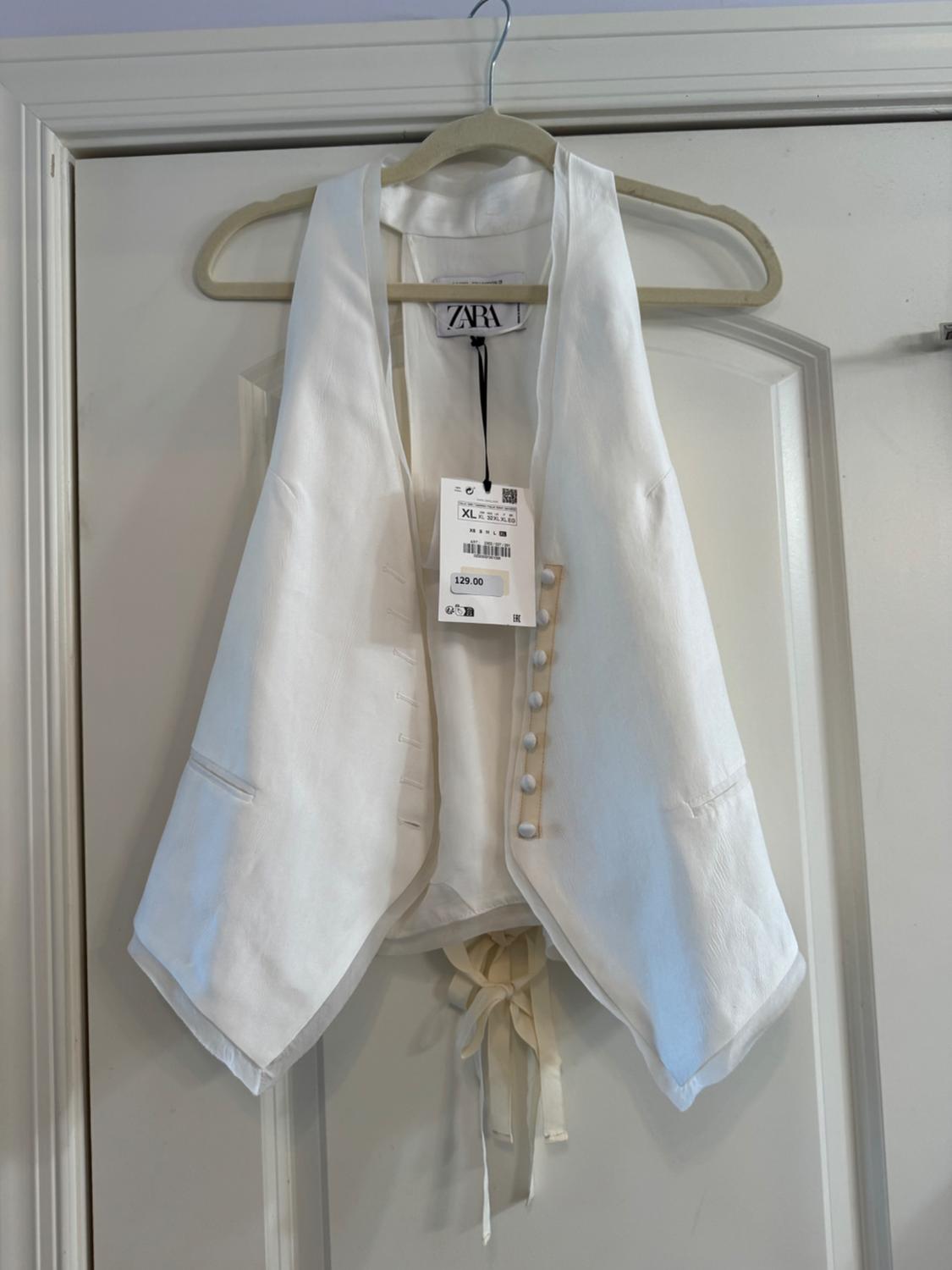 White vest hanging on a hanger with visible price tag of 129.00. White door background. ZARA brand on the label.