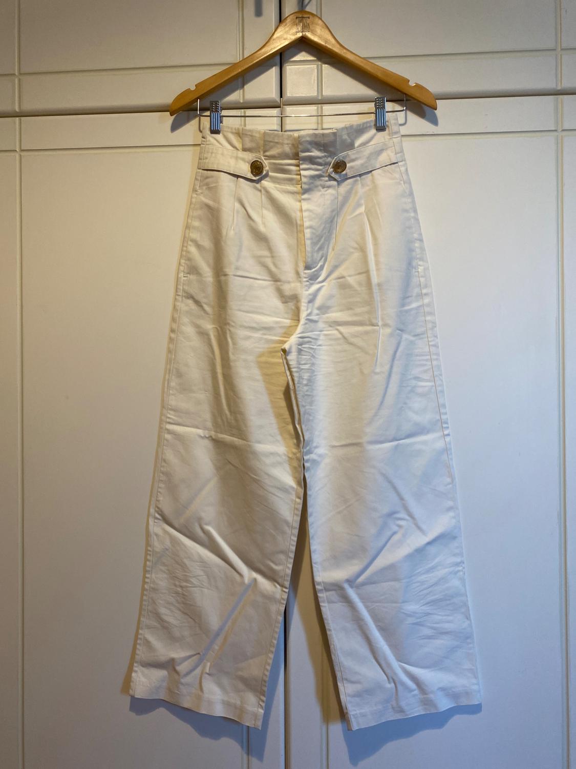 White wide-leg trousers hanging on a wooden hanger, with gold buttons at the waist, white wardrobe background.