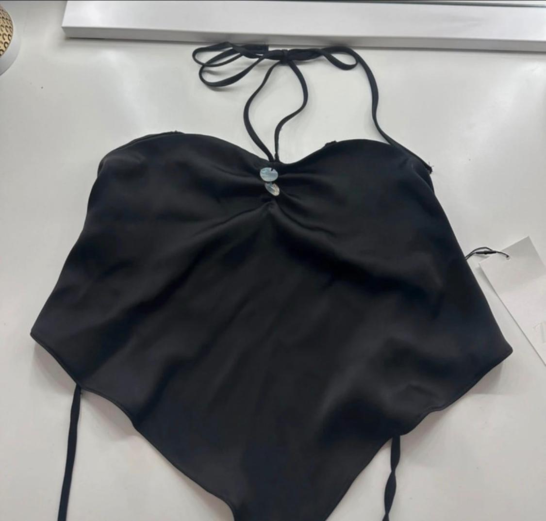Black halterneck crop top with thin straps, gathered detail with appliqués on the neckline and an asymmetrical pointed hem, on a white background.