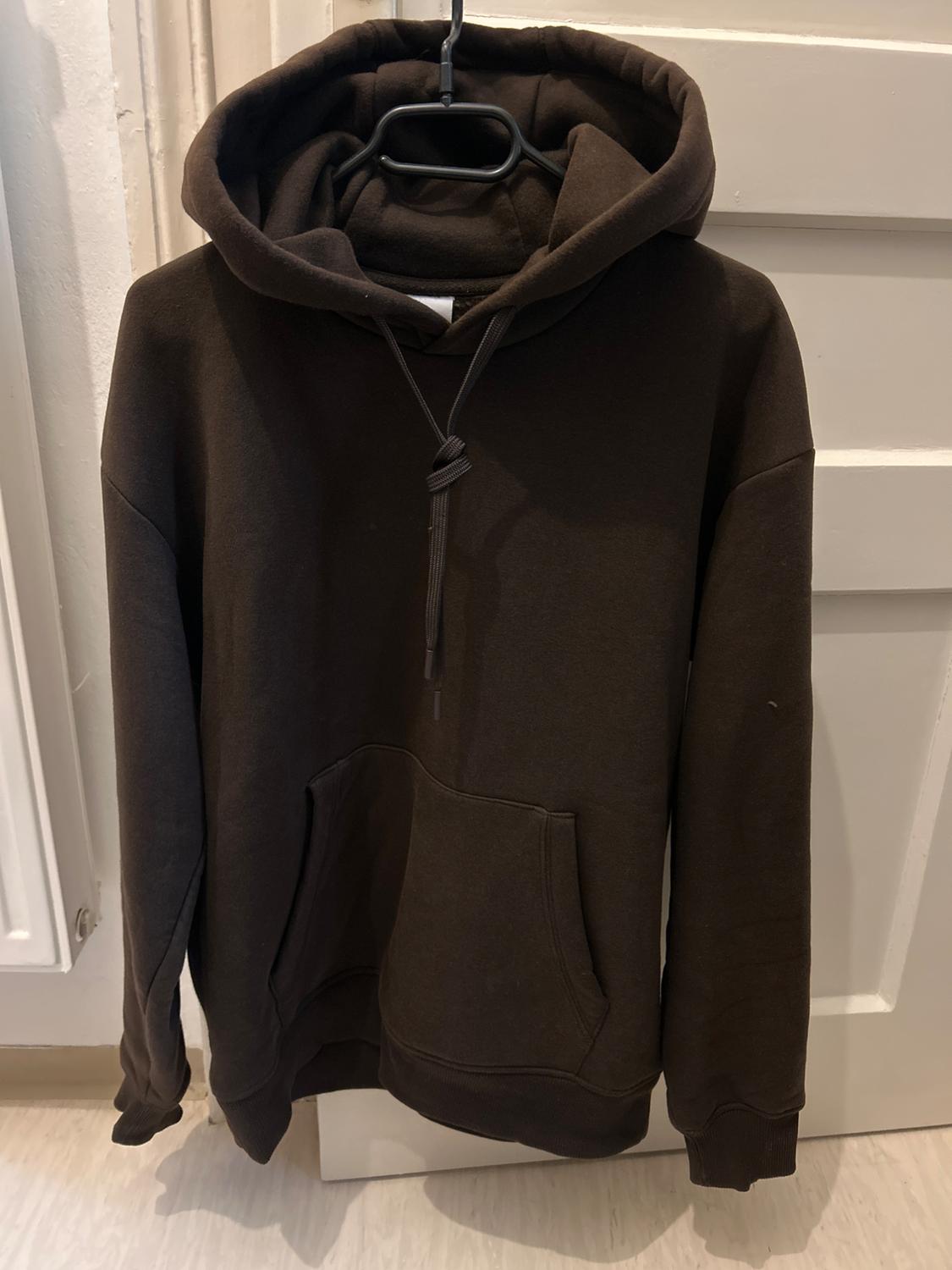 Brown hoodie with a front pocket, hanging on a black hanger in front of a white door.