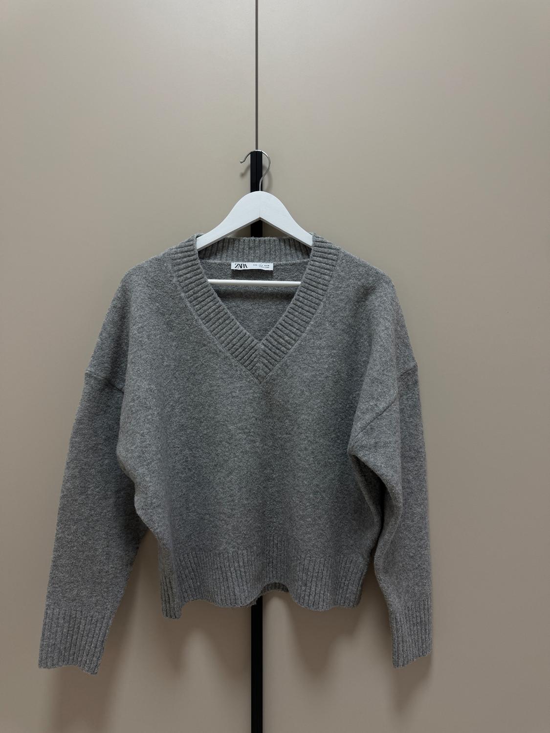 Grey knitted V-neck jumper hanging on a white hanger in front of a beige background. Visible label with text "ZARA".