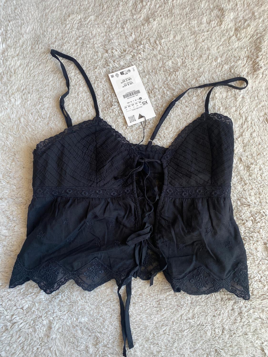 Black, thin-strapped camisole with lace and a front bow, on a beige fabric background. Visible label with size details and barcode.