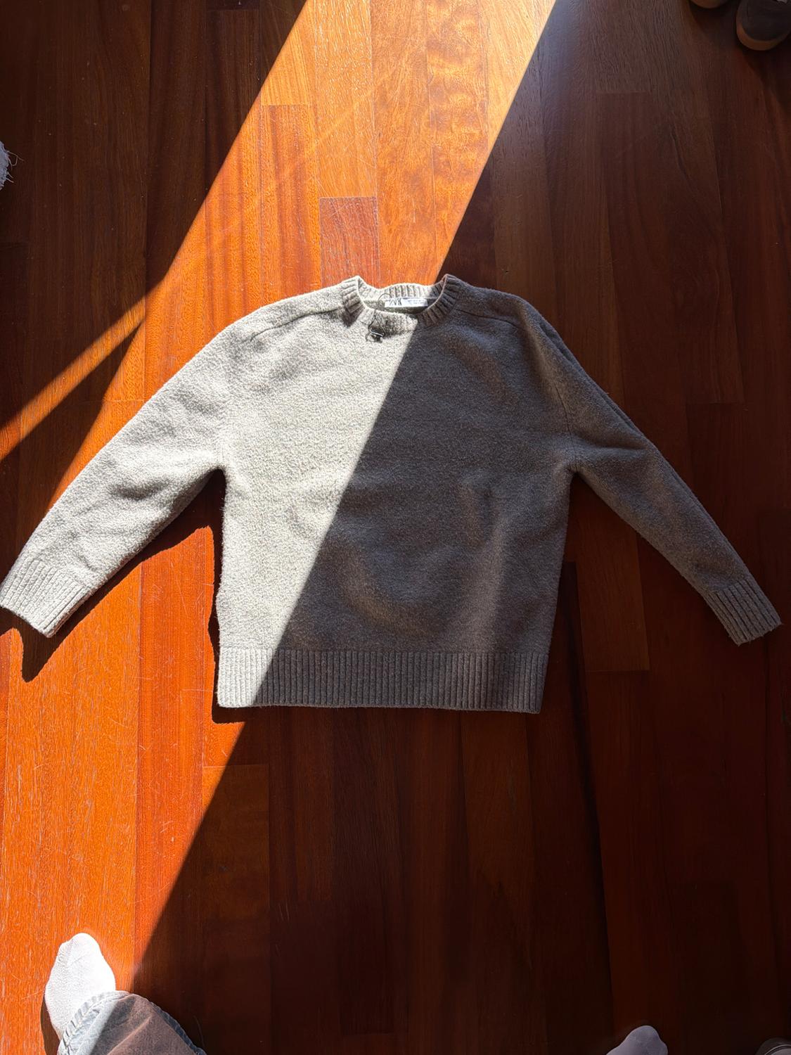 Grey long-sleeved jumper on a wooden floor lit by a sunbeam. Part of a foot with a white sock visible in the corner.